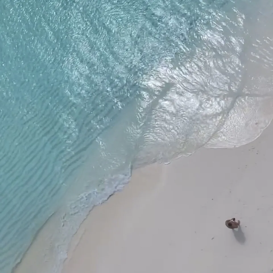 Looking straight down at the edge of the lagoon at Coco Palm Dhuni Kolhu, ripples in pale turquoise water running up onto white sand, one person standing at the waterline