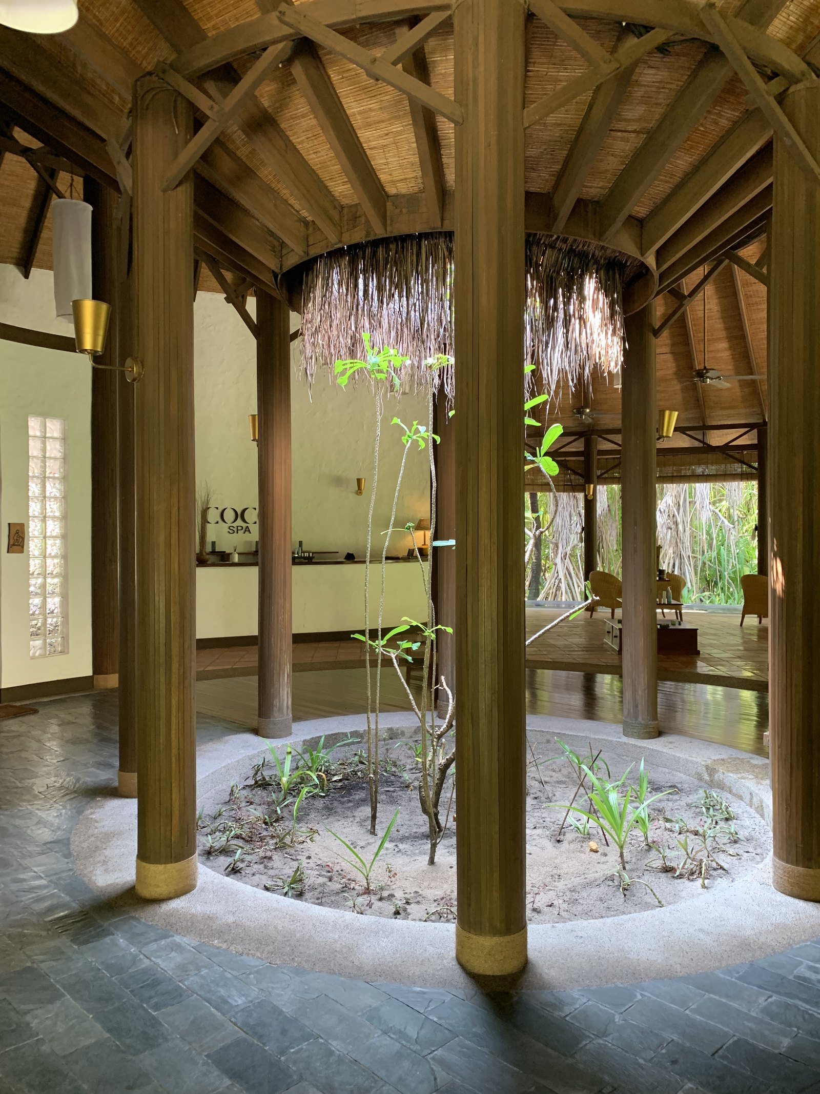 The timber colonnade of the spa at Coco Palm Dhuni Kolhu, posts and a thatched roof around a planted bed of sand and young shoots