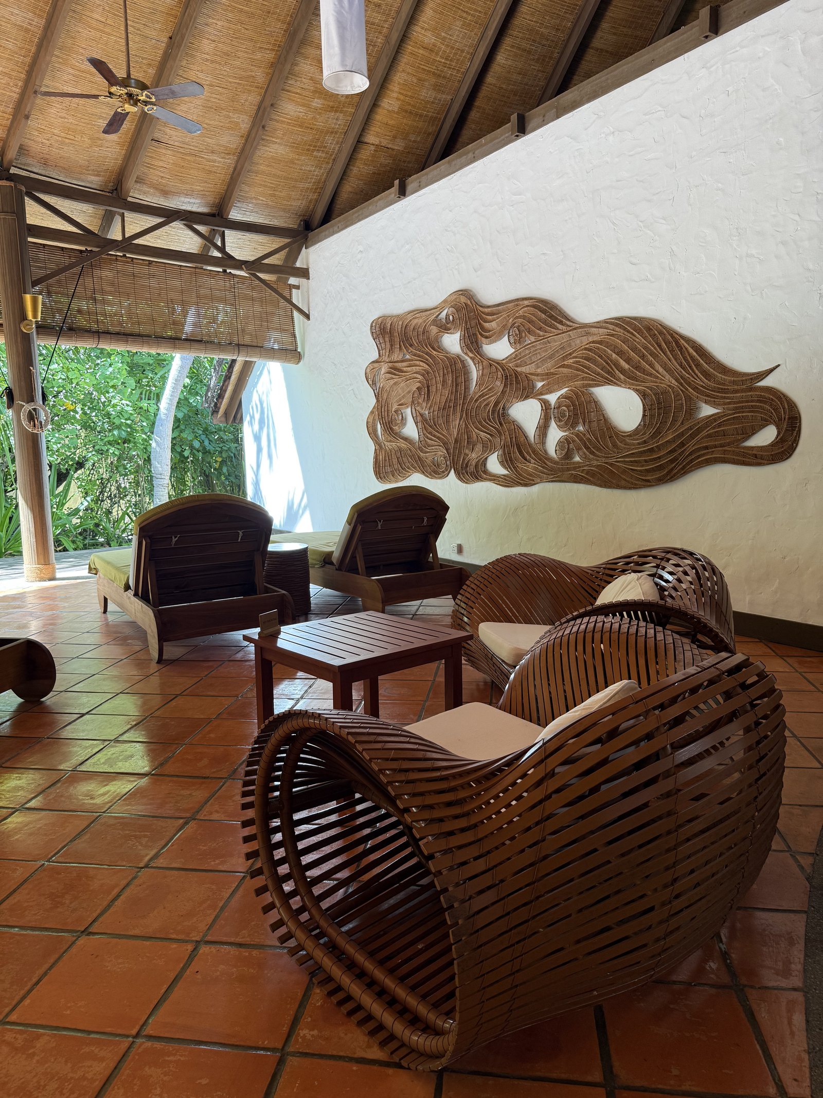 The relaxation room at the spa at Coco Palm Dhuni Kolhu, timber loungers and woven rattan chairs on a terracotta floor under a thatched roof, a long carved timber relief of a face on the white wall