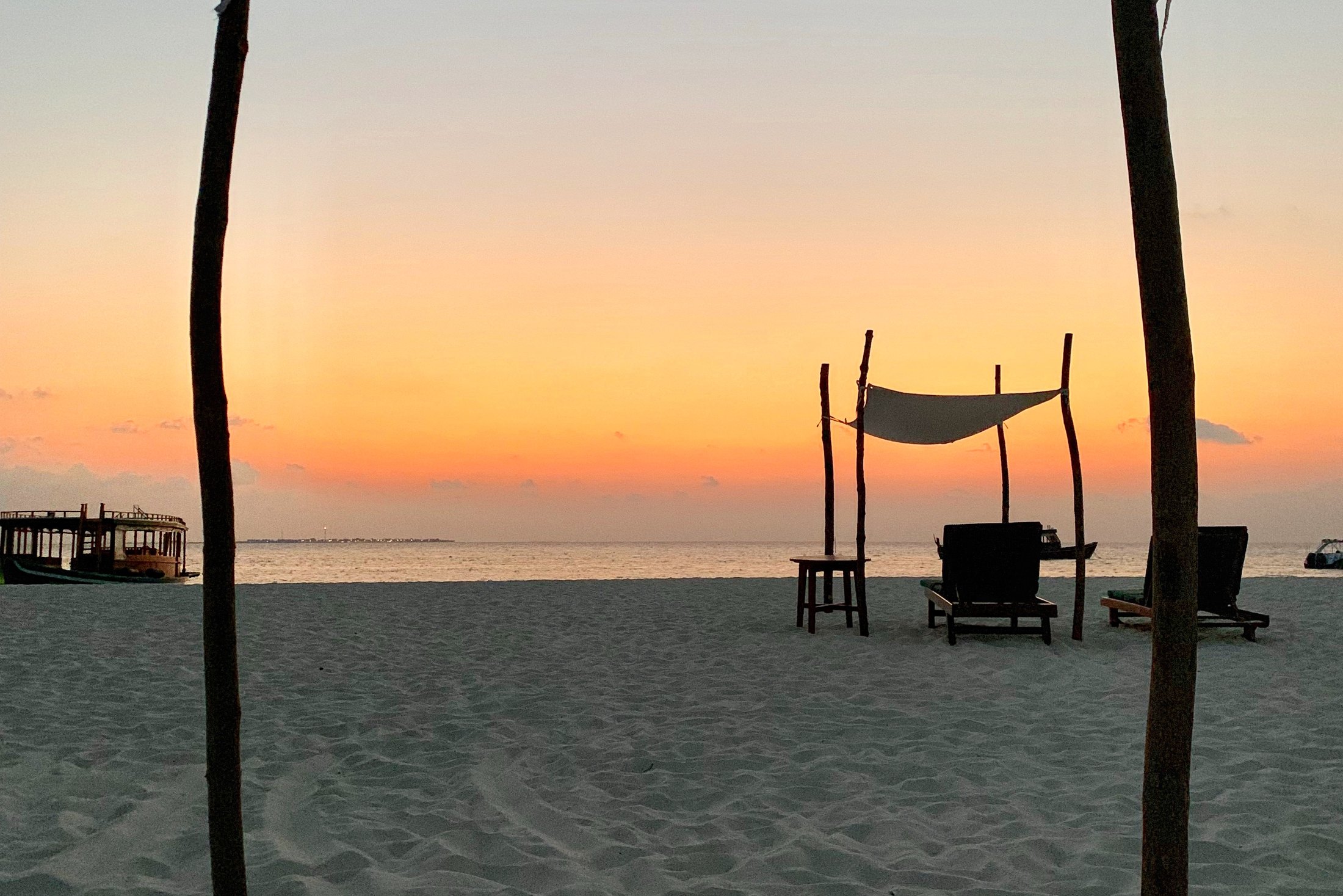 Sail canopies on timber poles over empty loungers on the beach at Coco Palm Dhuni Kolhu, the sun down and the sky orange over a flat sea