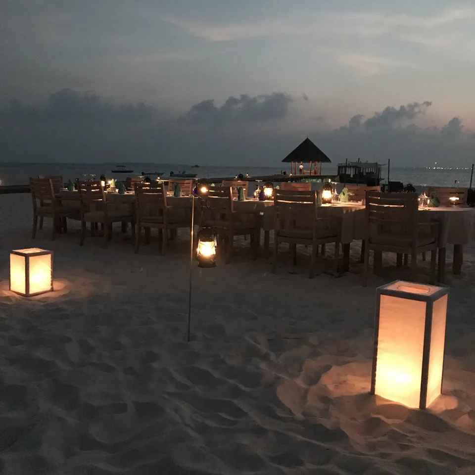 Long timber tables laid for dinner directly on the sand at dusk, lanterns burning on the ground between them and a thatched pavilion behind