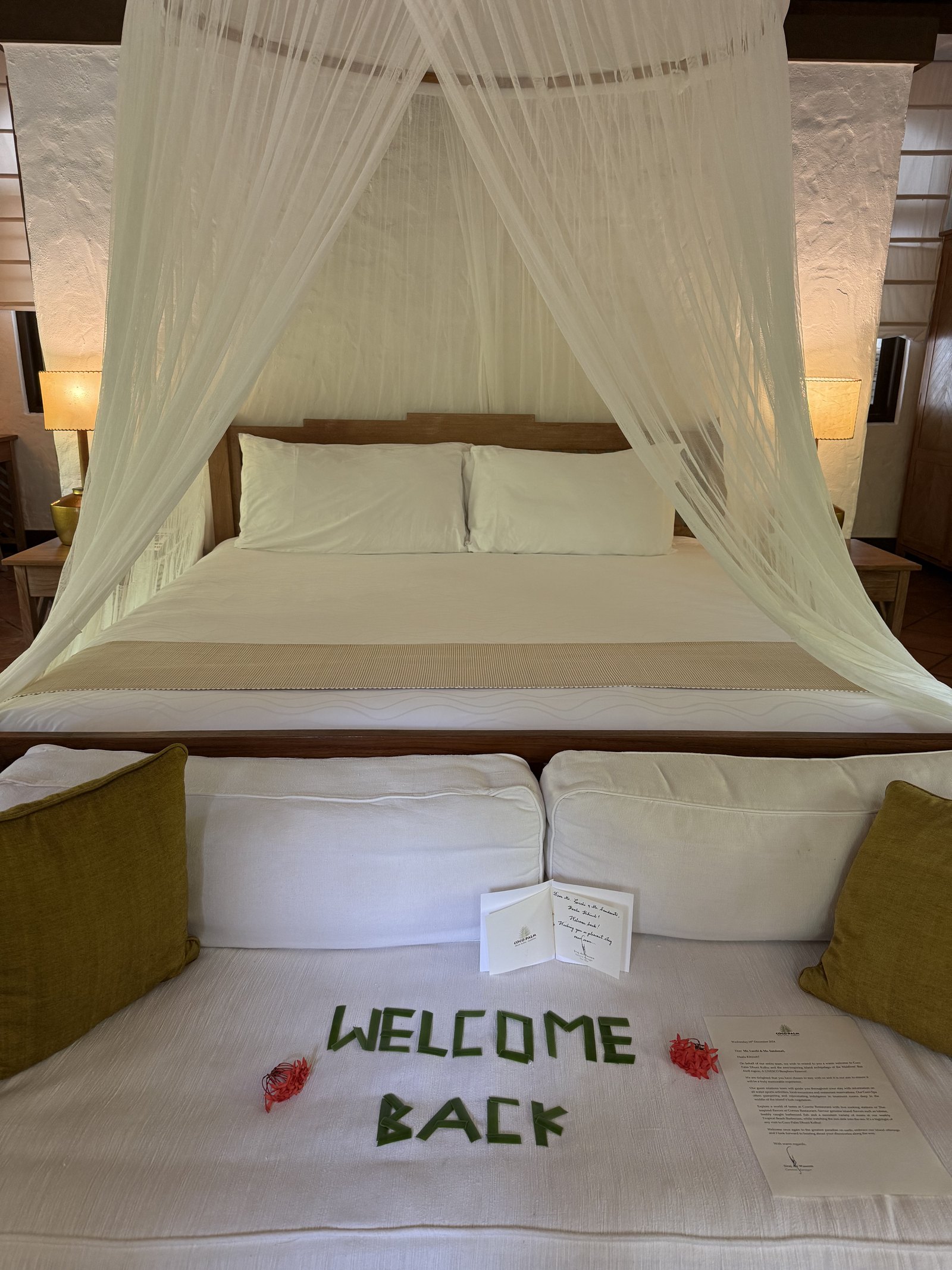 A bed at Coco Palm Dhuni Kolhu under a white net with WELCOME BACK spelled out in green leaves across the white cover