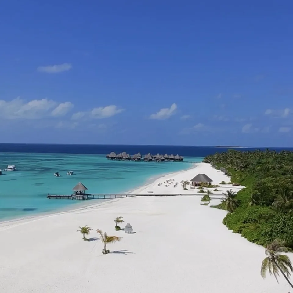 An aerial view of the wide white beach at Coco Palm Dhuni Kolhu, a thatched pavilion on the sand and a jetty running into the lagoon, the overwater villas along the reef edge beyond