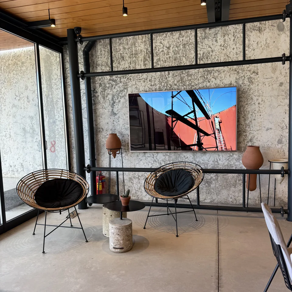 A framed photograph hung on a raw concrete wall in a lounge at Dexamenes, wire chairs and ceramic vessels in front of it under a timber ceiling