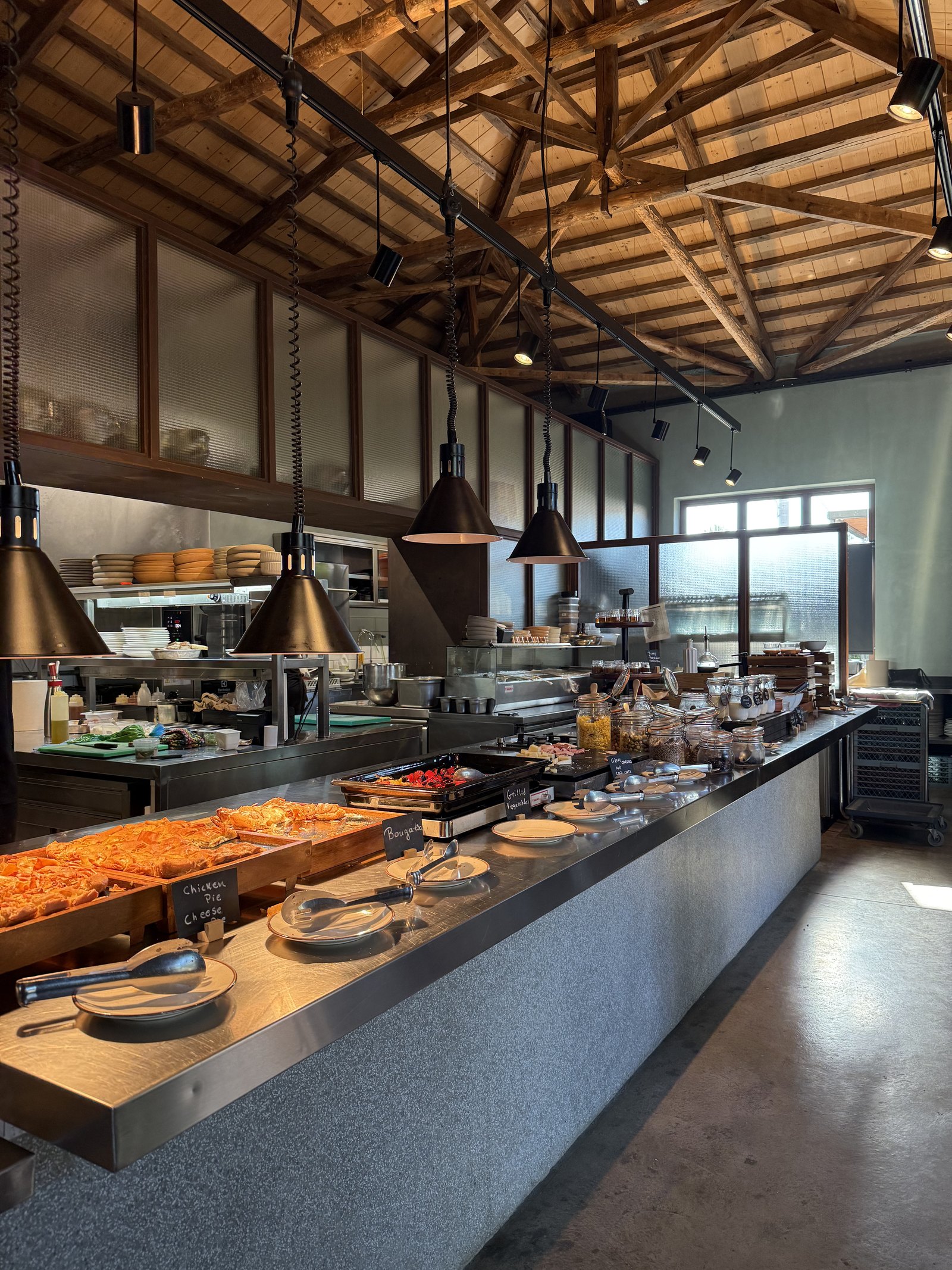 The breakfast counter at Dexamenes under an open timber roof structure, trays of food along a long steel top with brass lamps above