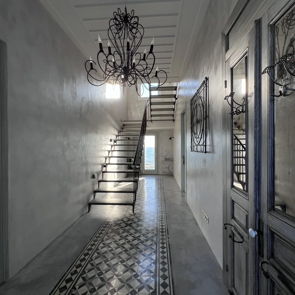 A narrow white corridor at Dexamenes with a patterned black and white tiled floor, an old iron chandelier and a steel stair at the end