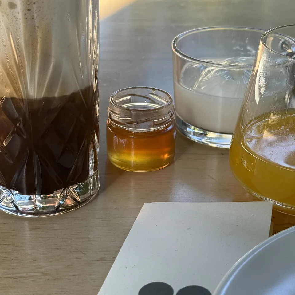A tall glass of freddo espresso beside a small jar of honey and a glass of juice on a pale timber table, a Dexamenes card alongside