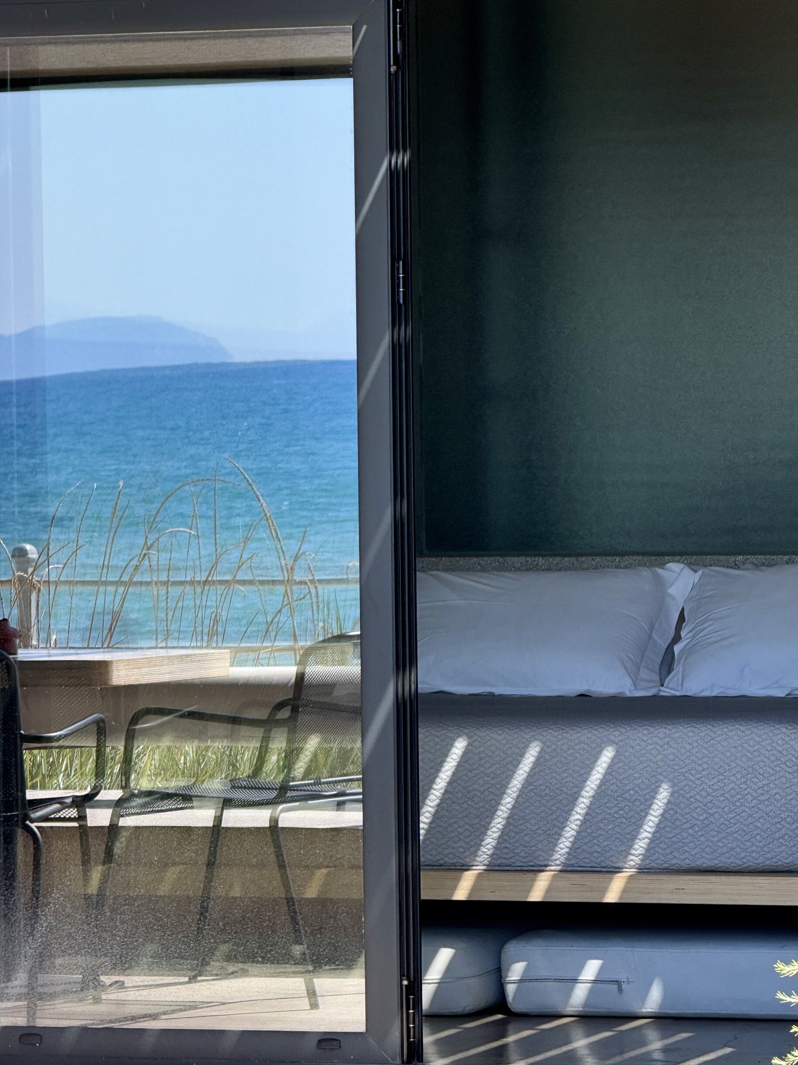 Looking out from inside a suite at Dexamenes through a sliding glass door past a low bed to the terrace and the blue sea beyond