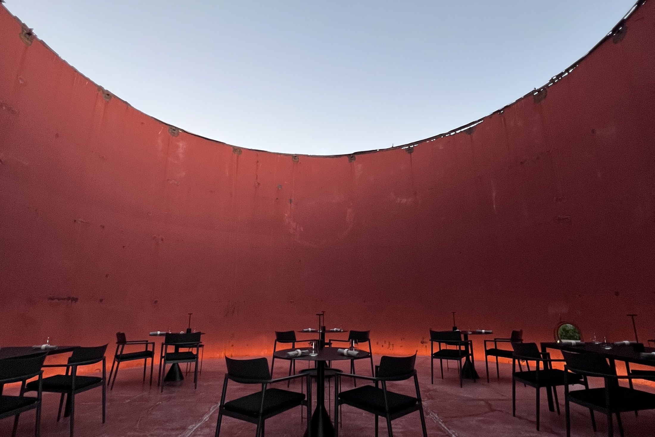 The inside of the 1920s silo at Dexamenes, a vast drum of oxide red concrete curving up to a perfect circle of open sky, with black tables and chairs set out on the floor