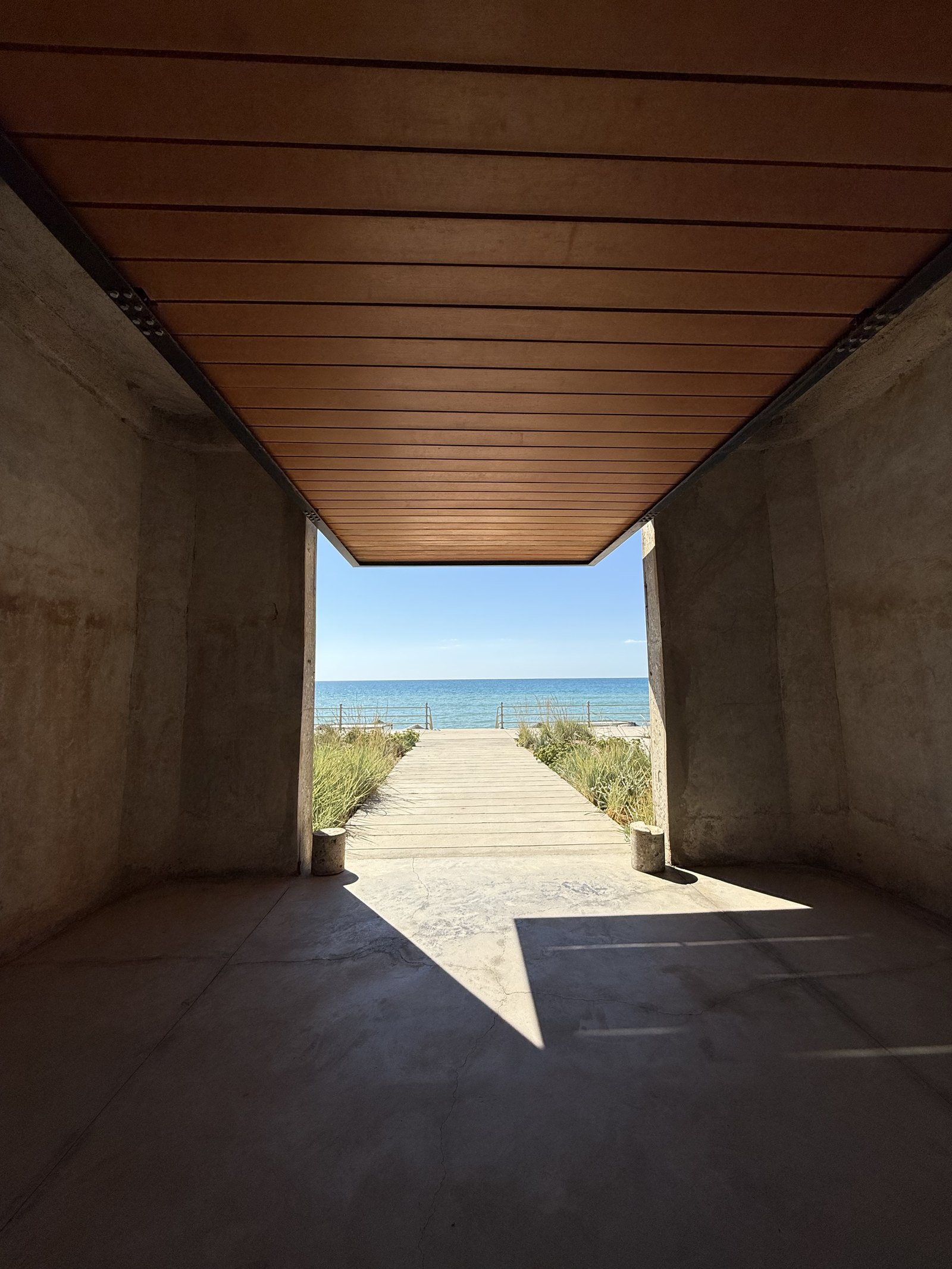 A raw concrete passage under a timber soffit at Dexamenes opening onto a boardwalk and the Ionian beyond