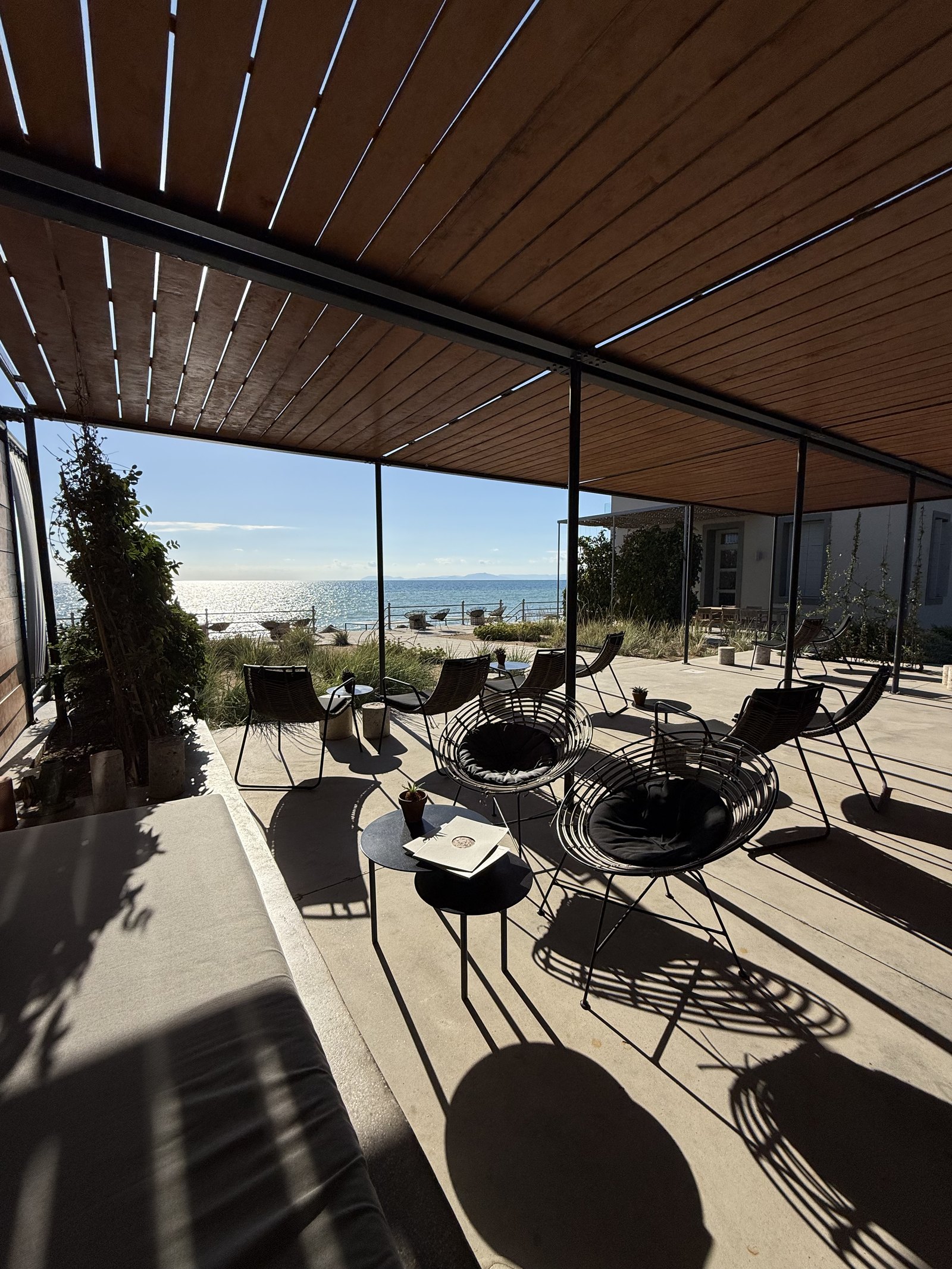 The long covered terrace at Dexamenes under a slatted timber pergola, wire chairs and low tables in a row and the sea at the far end