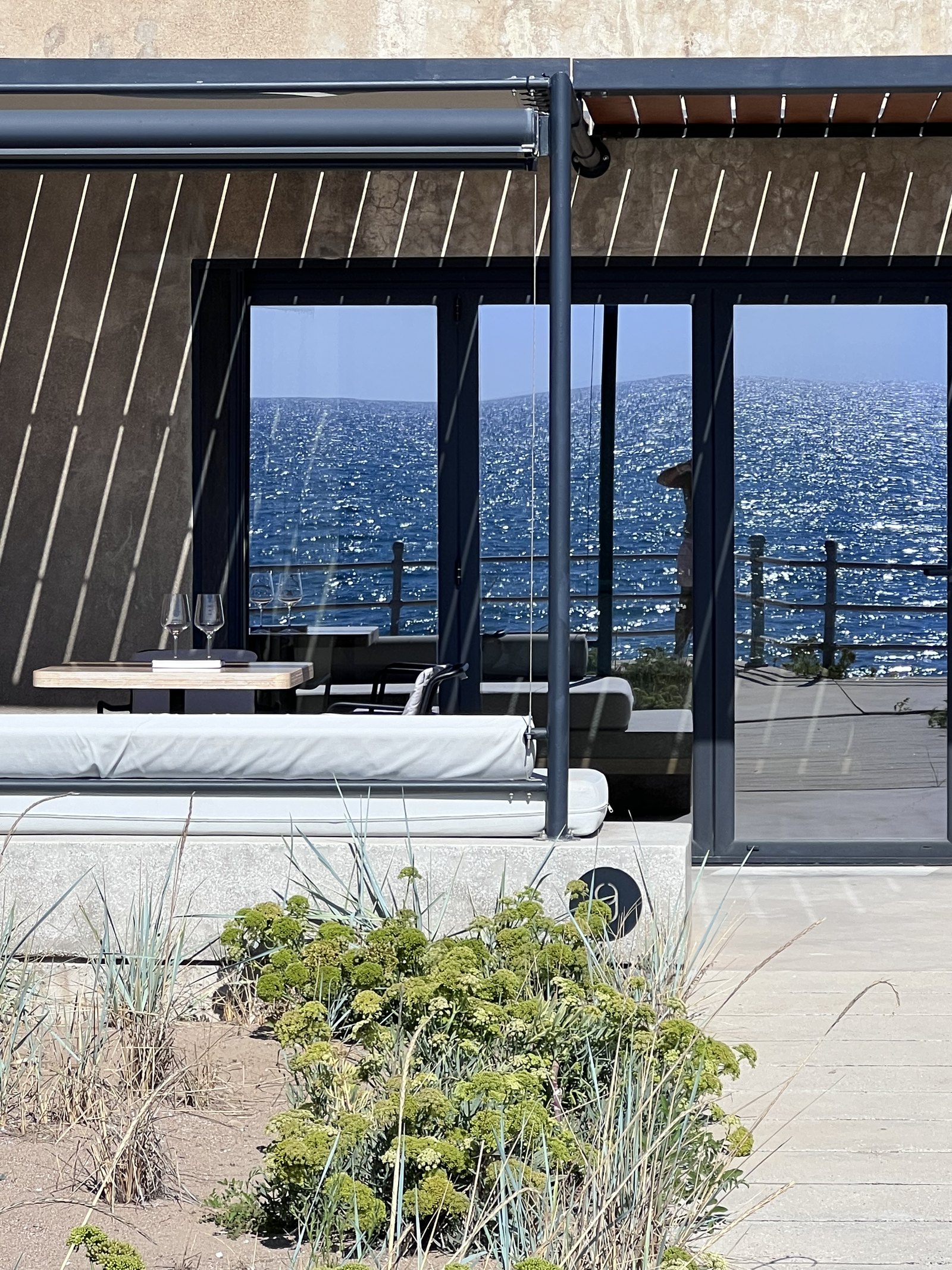 The screened patio of a beachfront suite at Dexamenes, a timber slat roof over a bench and table, with dune grass and the sea in front