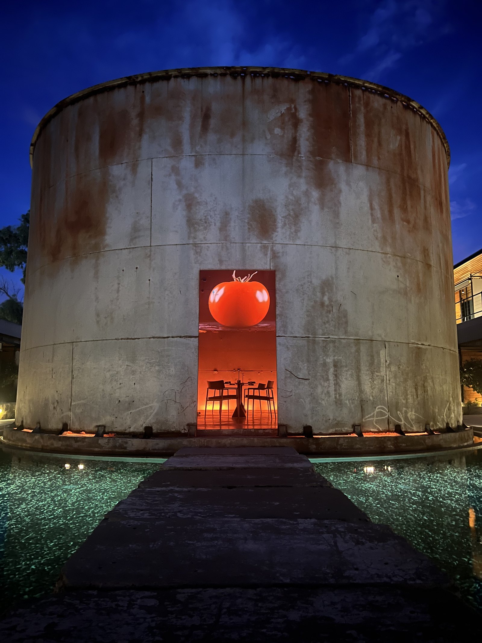 A 1920s concrete wine tank at Dexamenes lit at dusk, a tall red doorway cut into its curved wall with tables glowing inside and a shallow lit pool across the front