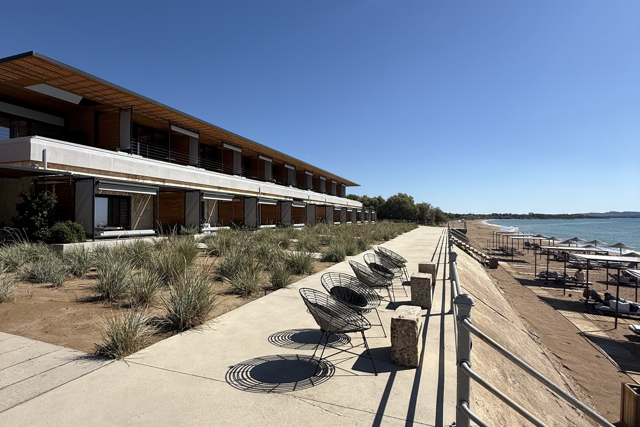 The long low line of suites at Dexamenes running down the beach behind a concrete terrace, wire chairs casting shadows, the sand and the Ionian at the end of it