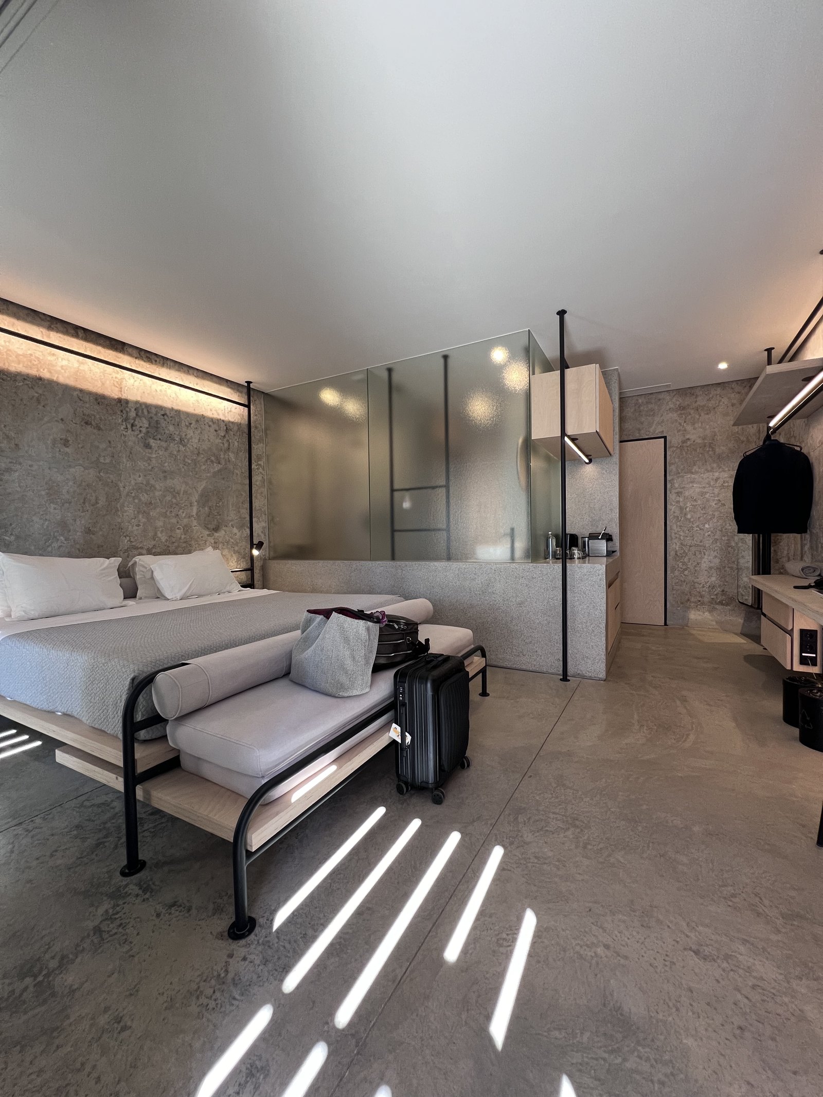 The inside of a tank suite at Dexamenes, a low bed and bench on black steel frames, raw concrete walls and bands of sunlight falling across the floor through a timber screen