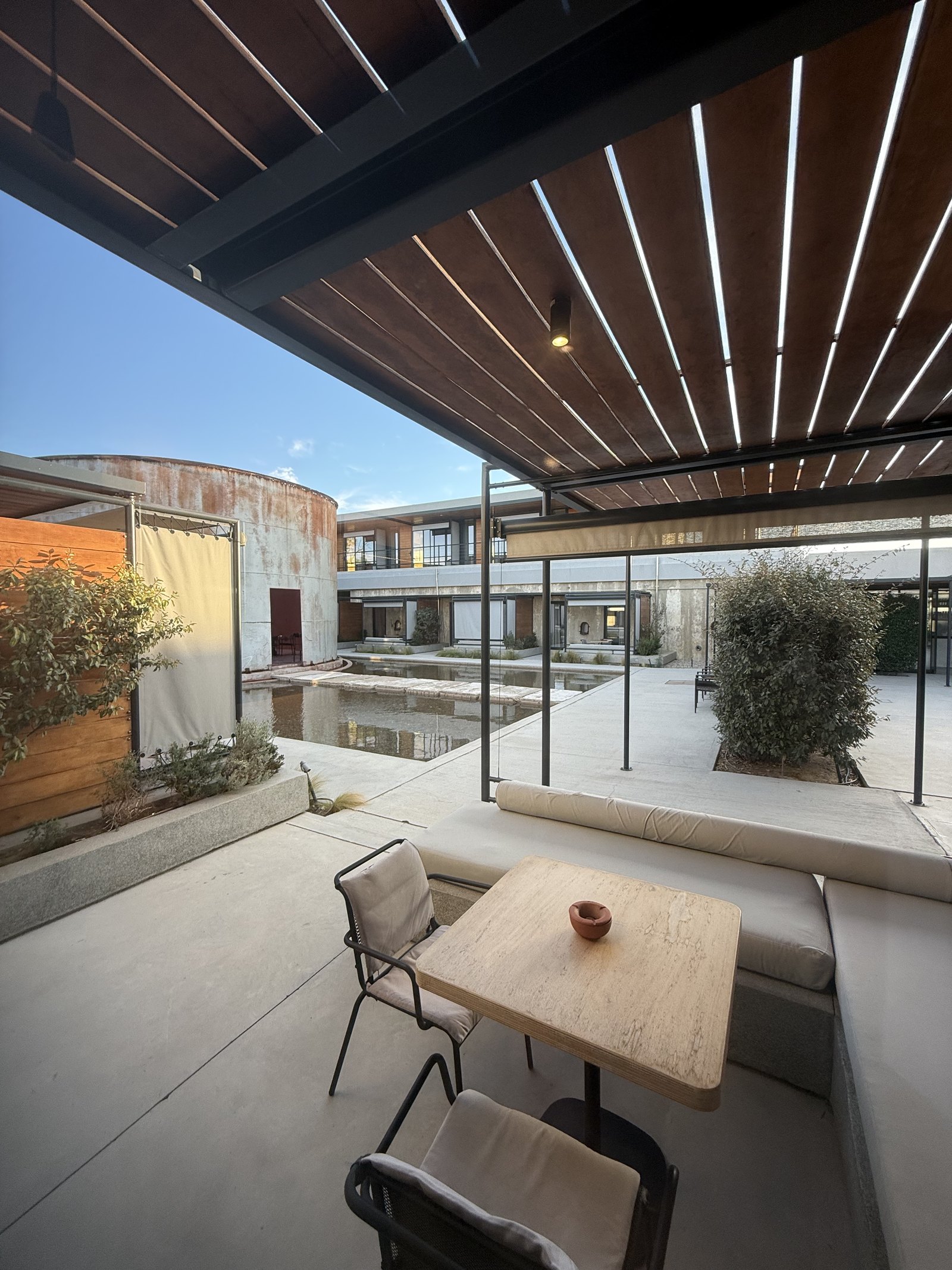 A terrace at Dexamenes with a small table and bench looking over a long shallow reflecting pool to a tank and the low suites beyond