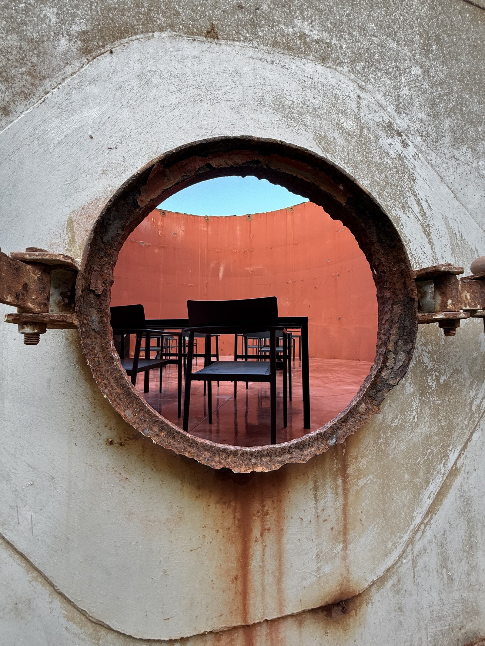 A rusted steel manhole ring bolted into the concrete wall of a tank at Dexamenes, looking through the opening to black chairs and tables in the red interior