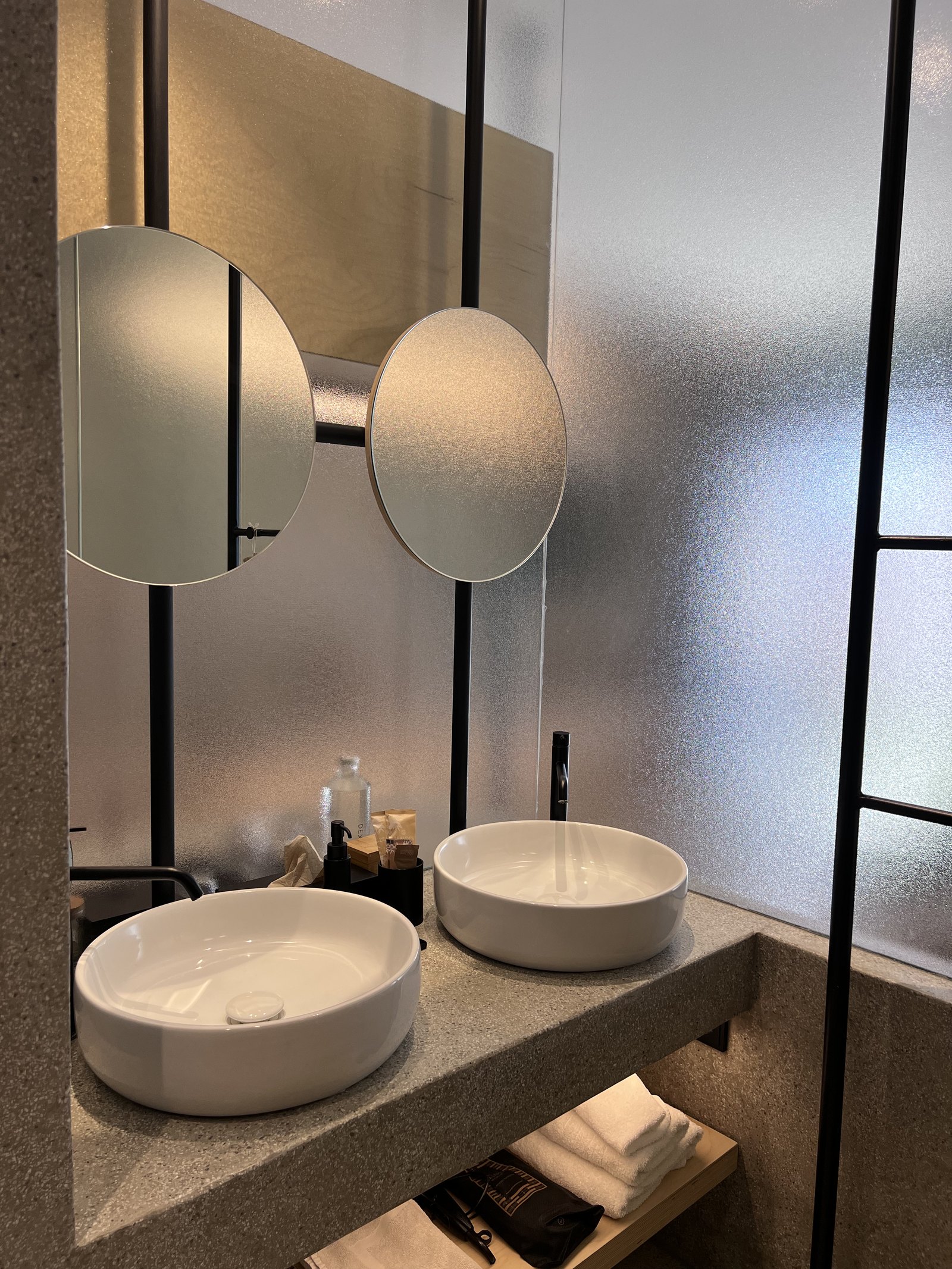 Two round white basins on a terrazzo shelf at Dexamenes under two round mirrors on black steel arms