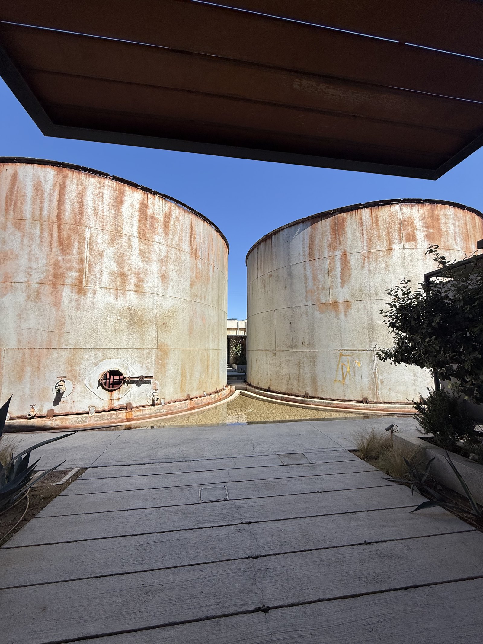 Two rusted concrete storage tanks standing side by side at Dexamenes with a timber boardwalk running between them and a heavy pipe at their base