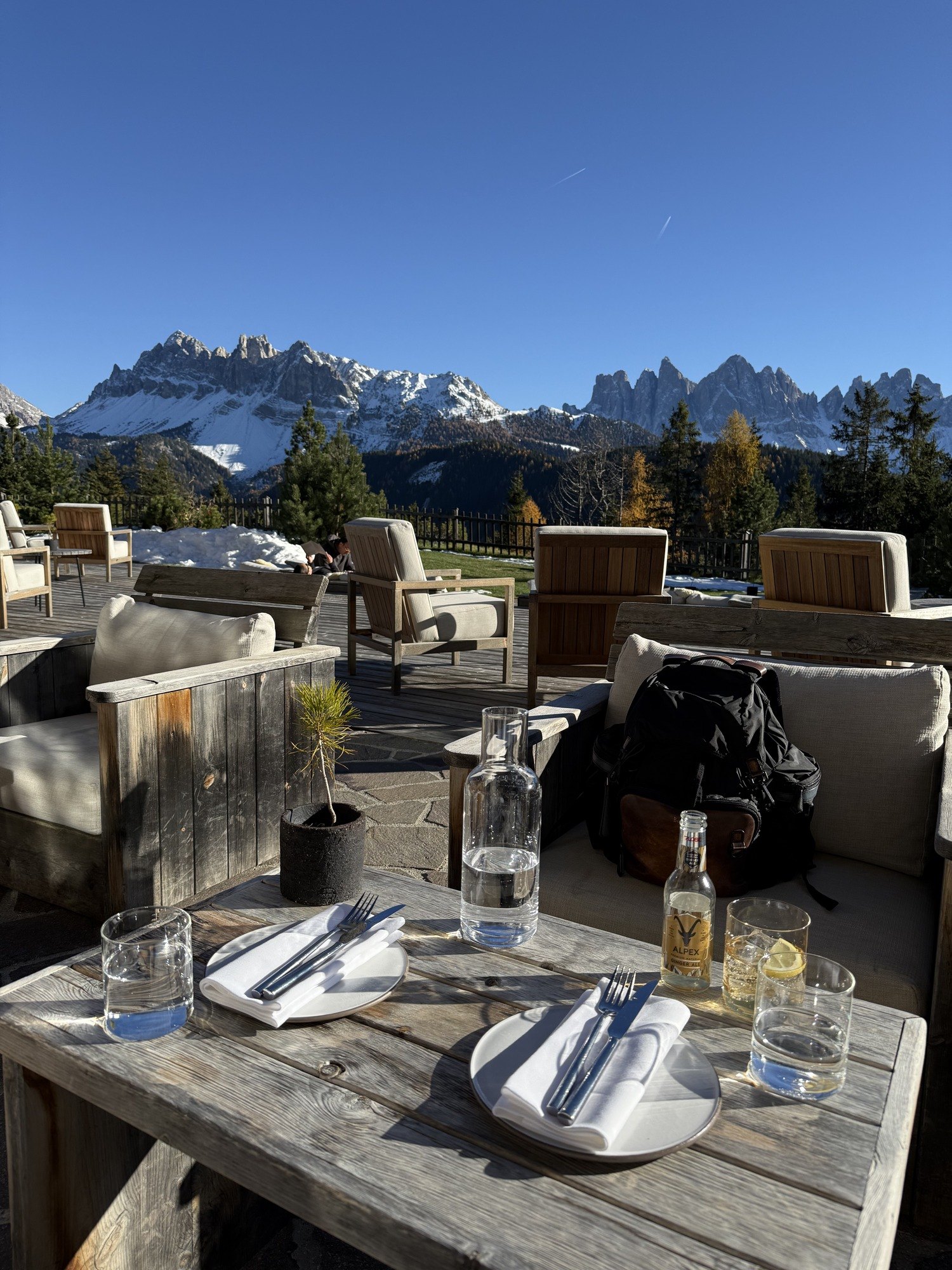 A weathered timber table on the terrace at FORESTIS laid with glasses and water, a rucksack on the bench and the Odle beyond