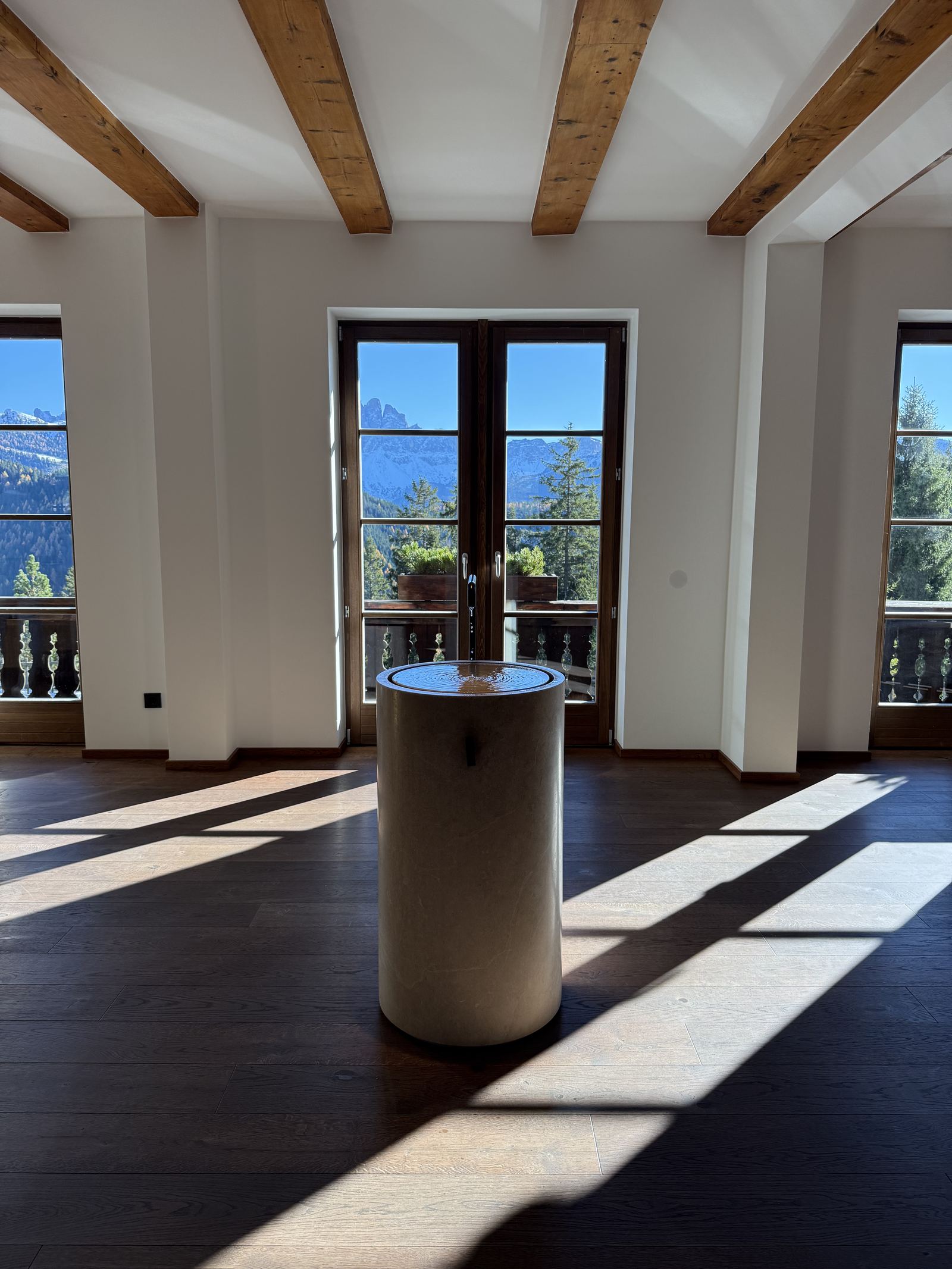A stone drinking fountain standing alone in an empty room at FORESTIS, with the Odle through the windows and bars of sunlight across the dark floor