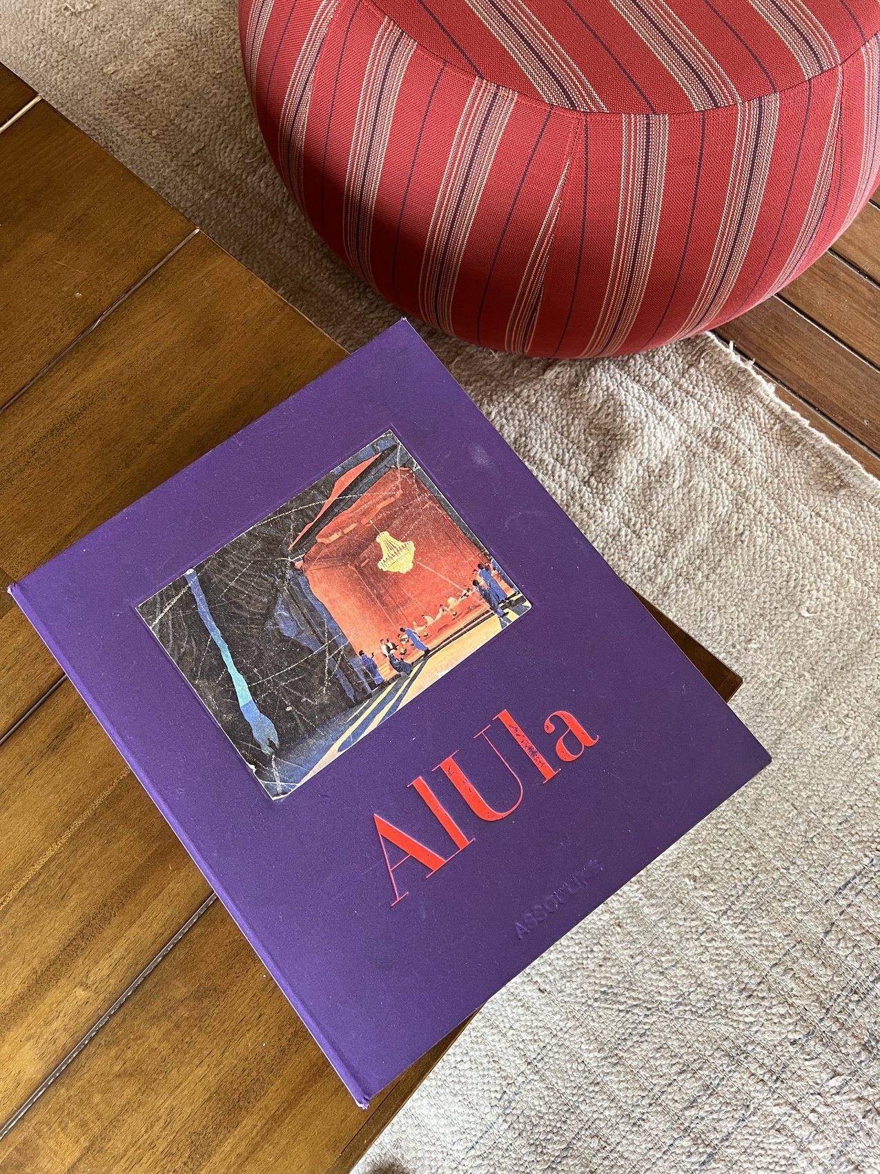 A purple book titled AlUla lying open on a woven rug beside a red striped cushion