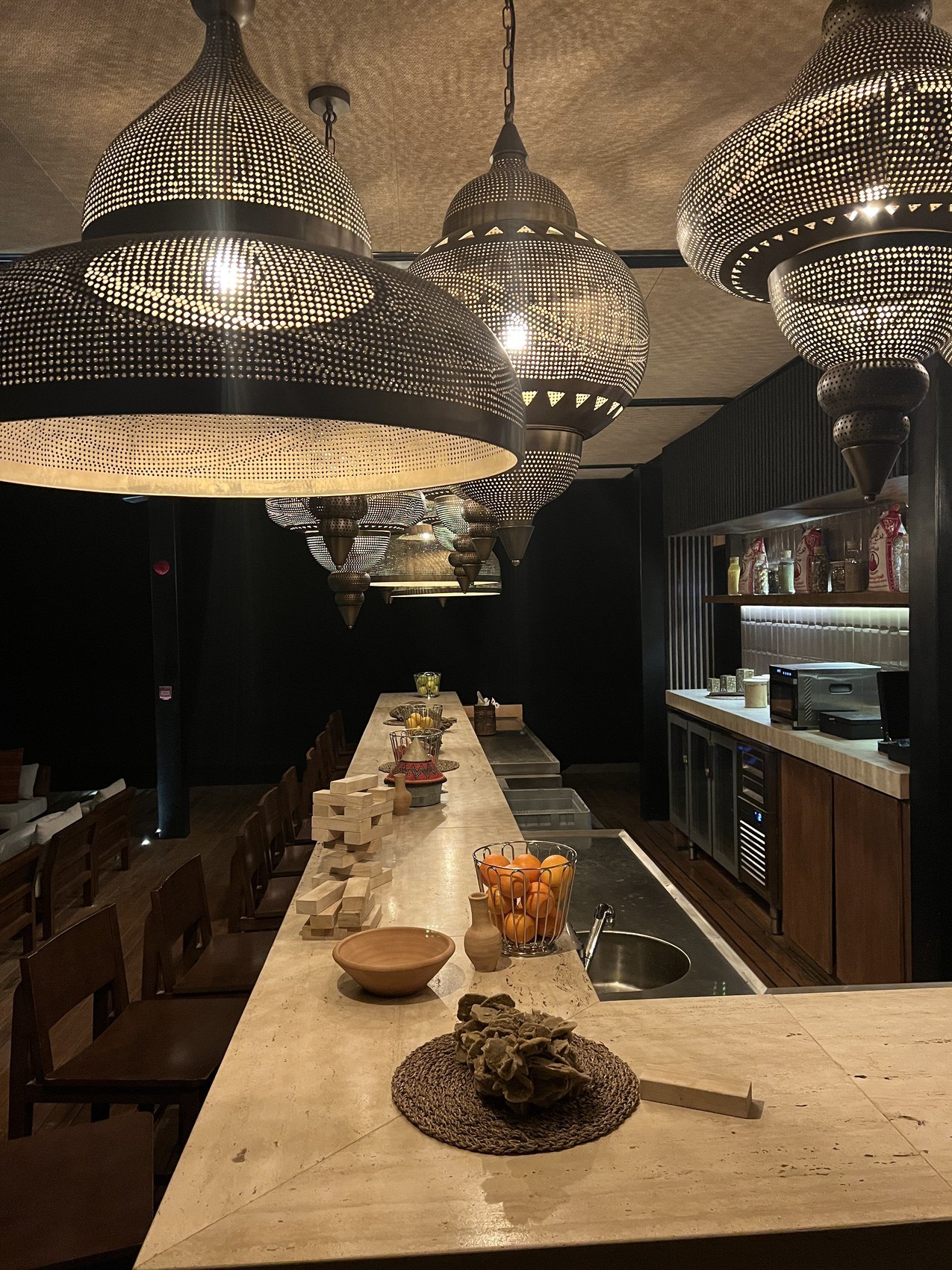 A long timber and marble counter at Habitas AlUla under perforated metal pendants with bottles and glassware behind
