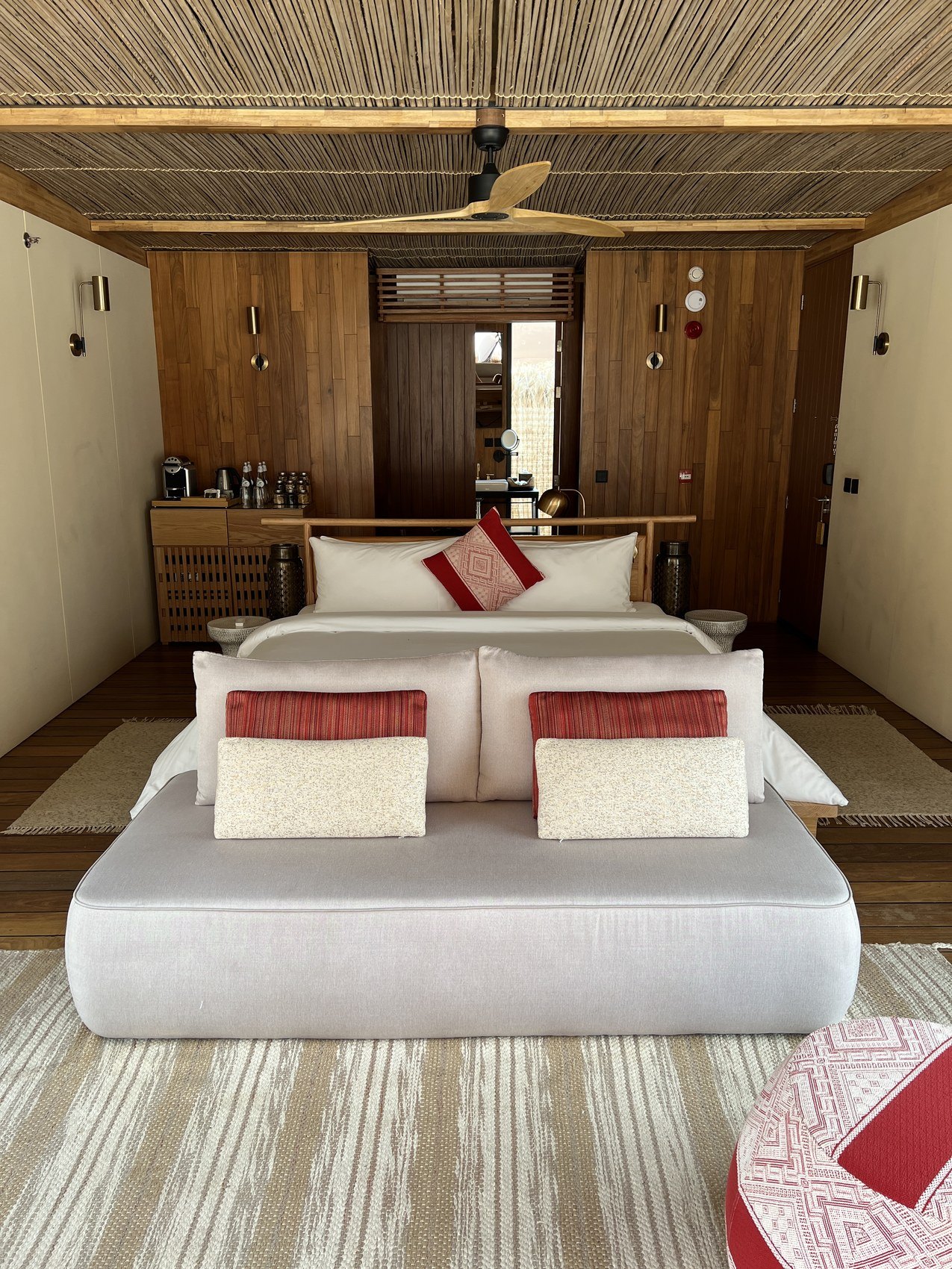 A low bed at Habitas AlUla with white linen and red striped cushions under a woven reed ceiling