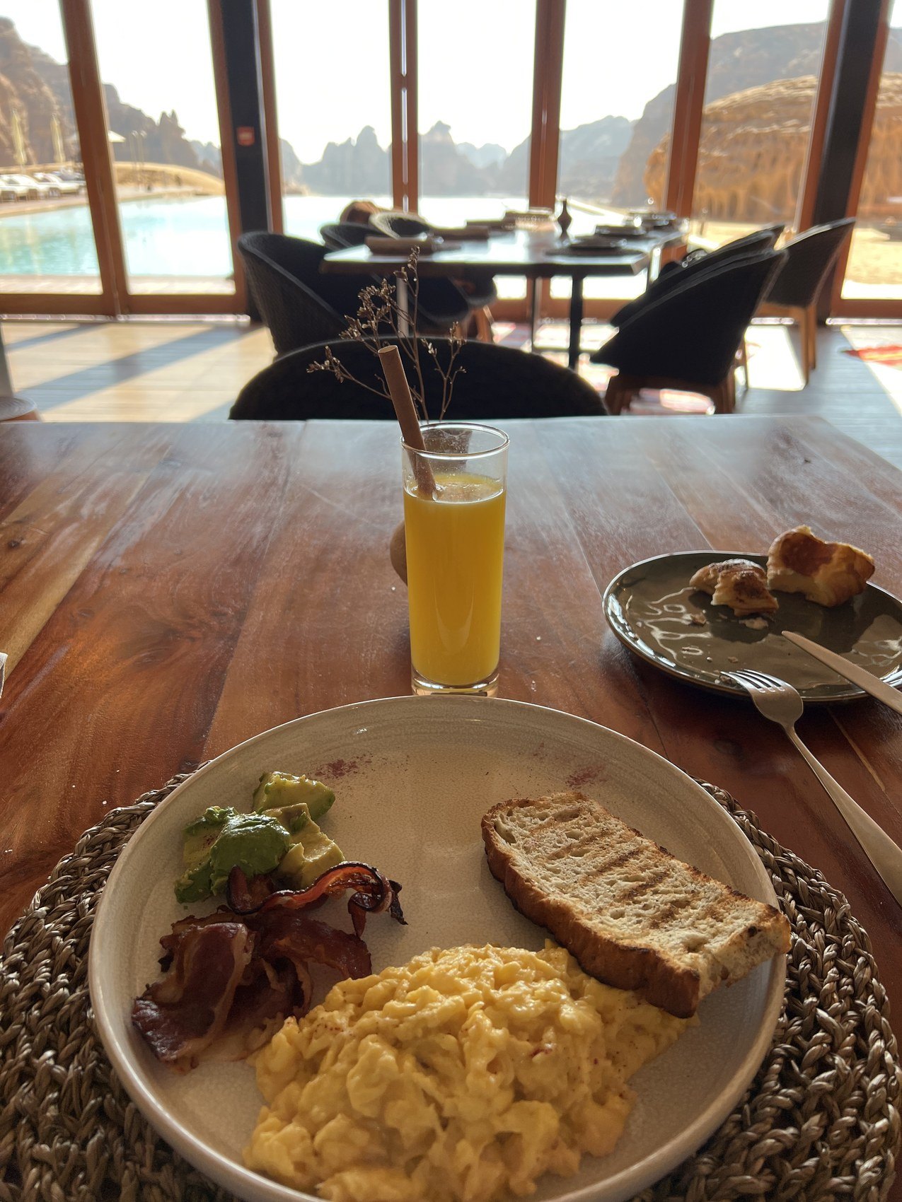 A breakfast plate with eggs, bacon and bread on a timber table at Habitas AlUla with the pool and the canyon through the glass behind