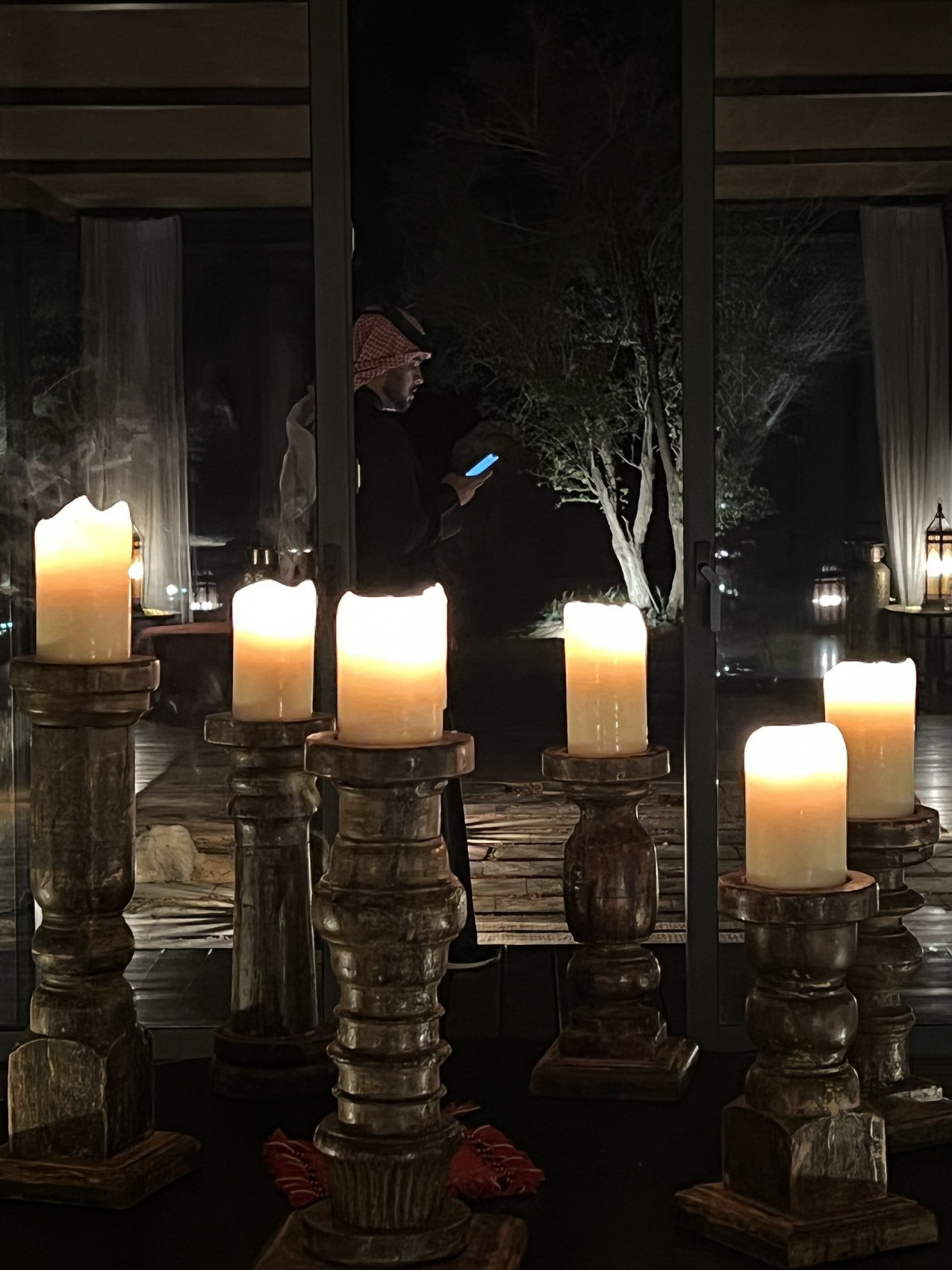 Thick candles burning on carved timber stands at night with glass and a dark room behind