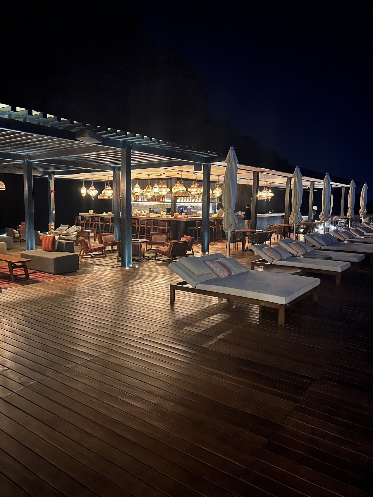 The pool deck at Habitas AlUla at dusk with loungers and closed parasols along the timber boards and the cliffs going dark behind