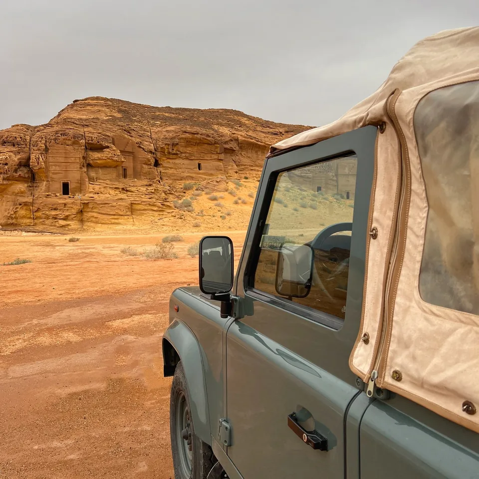 A vintage Land Rover Defender with a canvas top parked against the red rock at Hegra