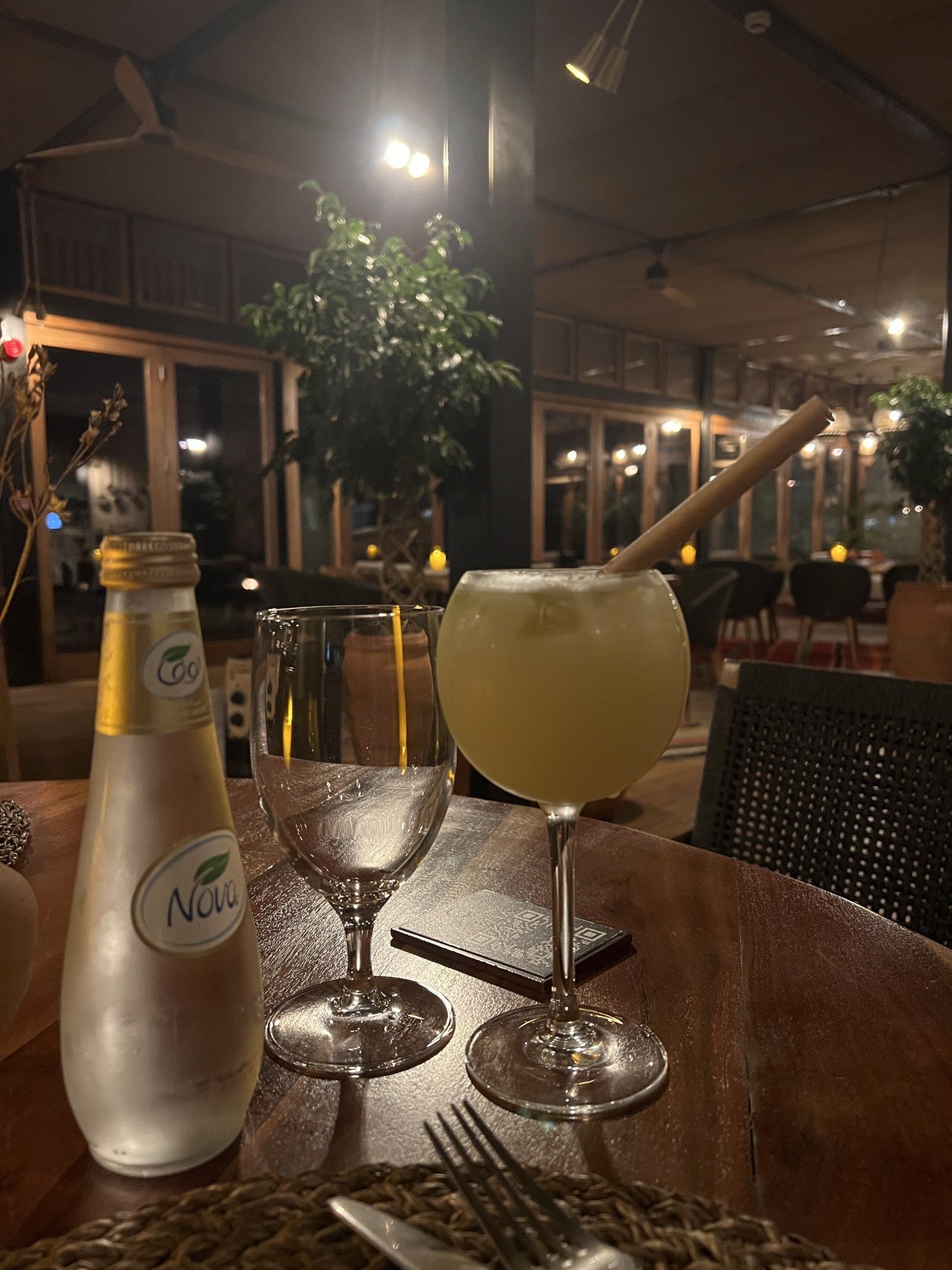 A glass of juice and a bottle of water on a table at Habitas AlUla in the evening with the lit lounge behind