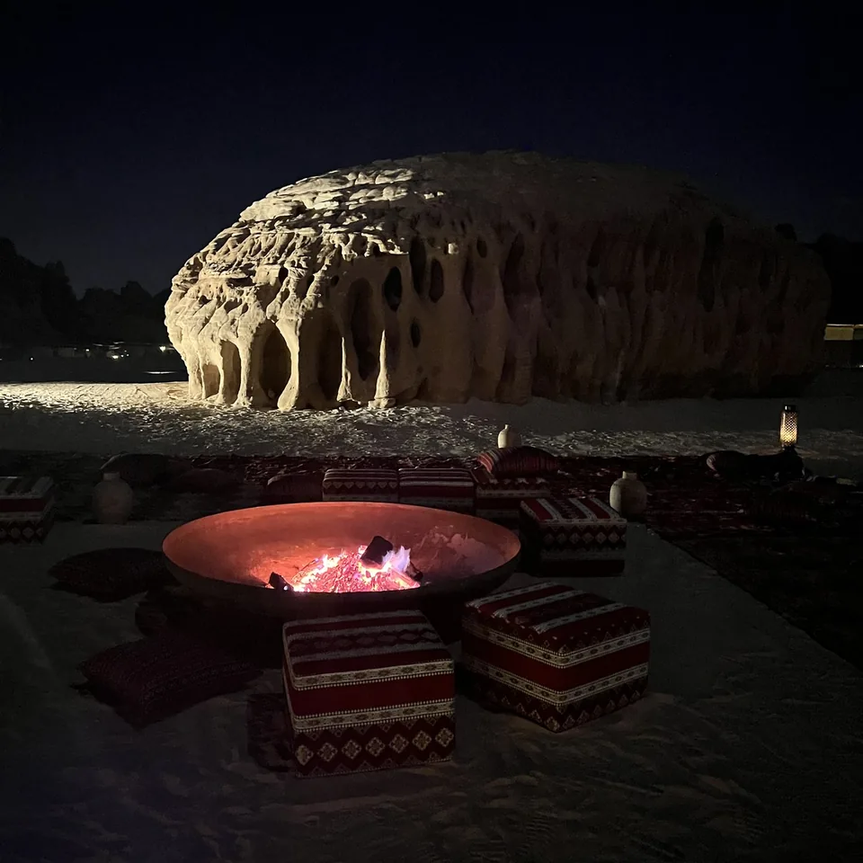 A round fire pit burning on the sand with the floodlit rock formation behind it