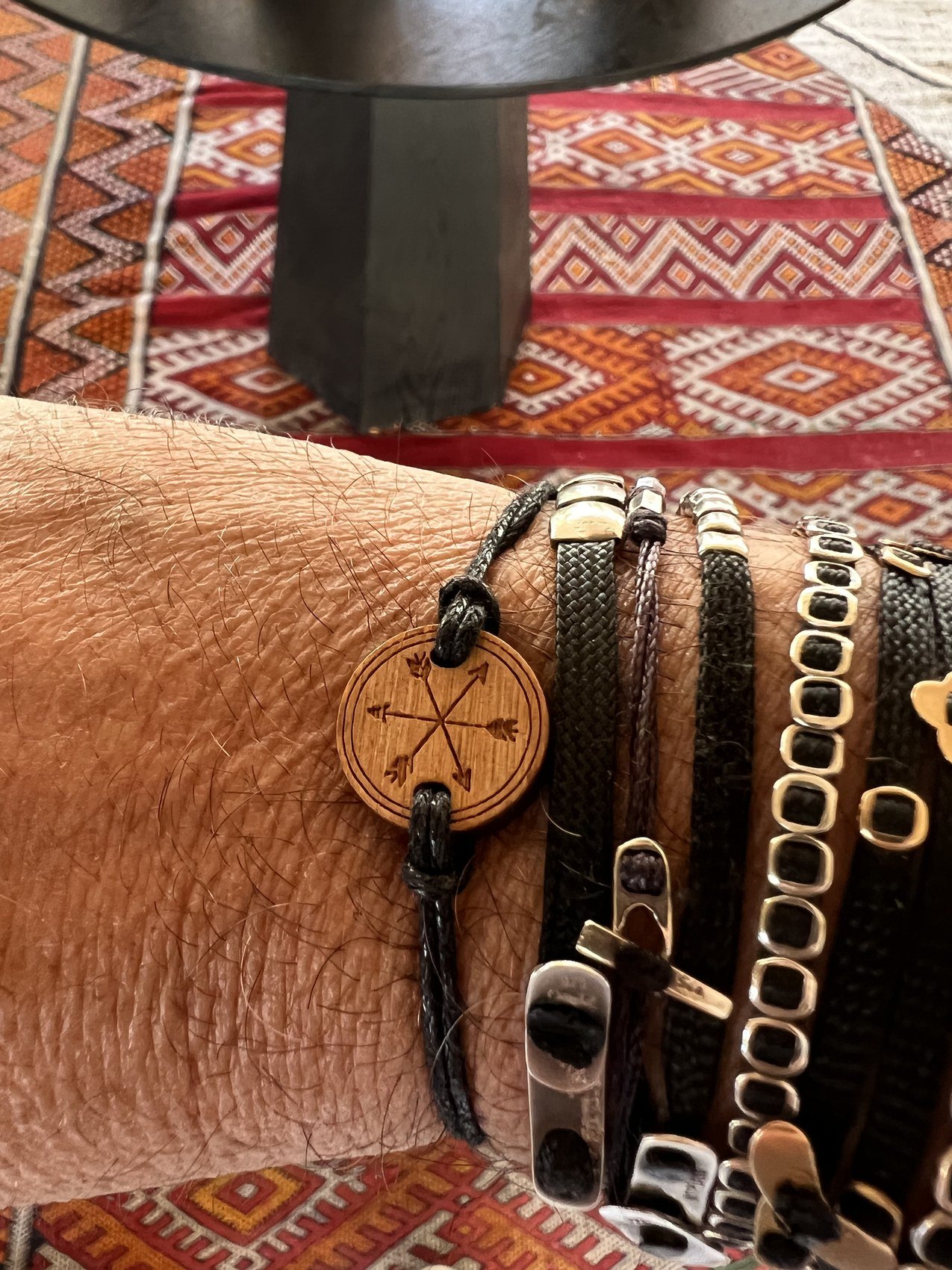 A wrist wearing several bracelets including a round wooden Habitas token, over a patterned kilim