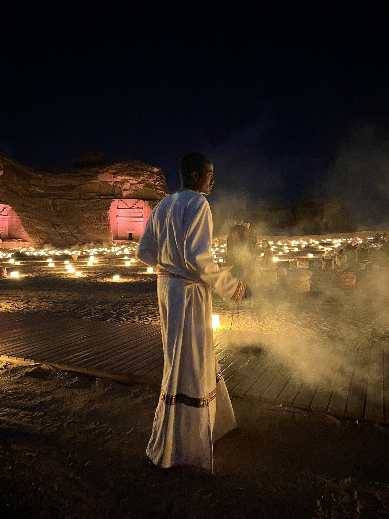 A figure in a white thobe carrying incense through the smoke at dusk above a terrace lined with candles