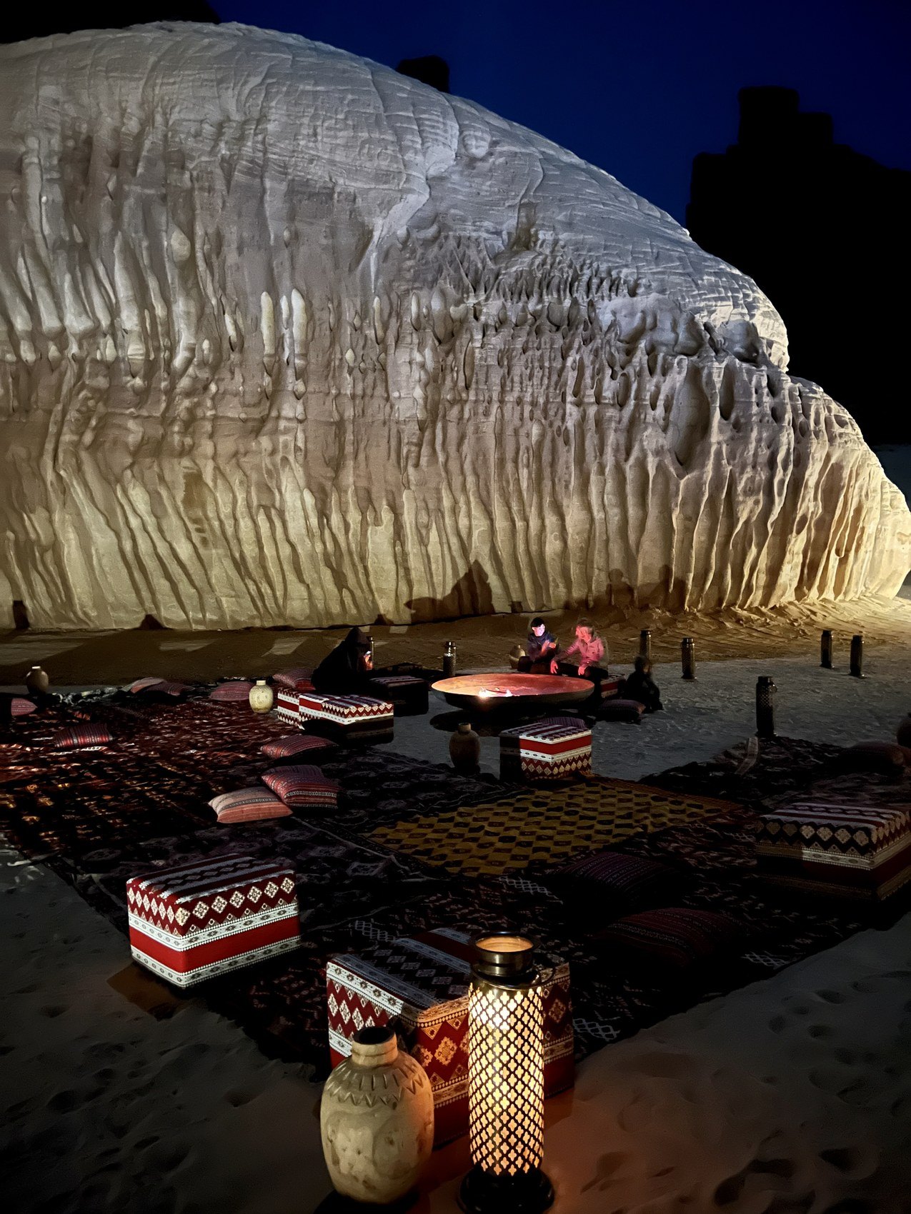 A floodlit sandstone formation at night above cushions, kilims and lanterns laid out on the sand