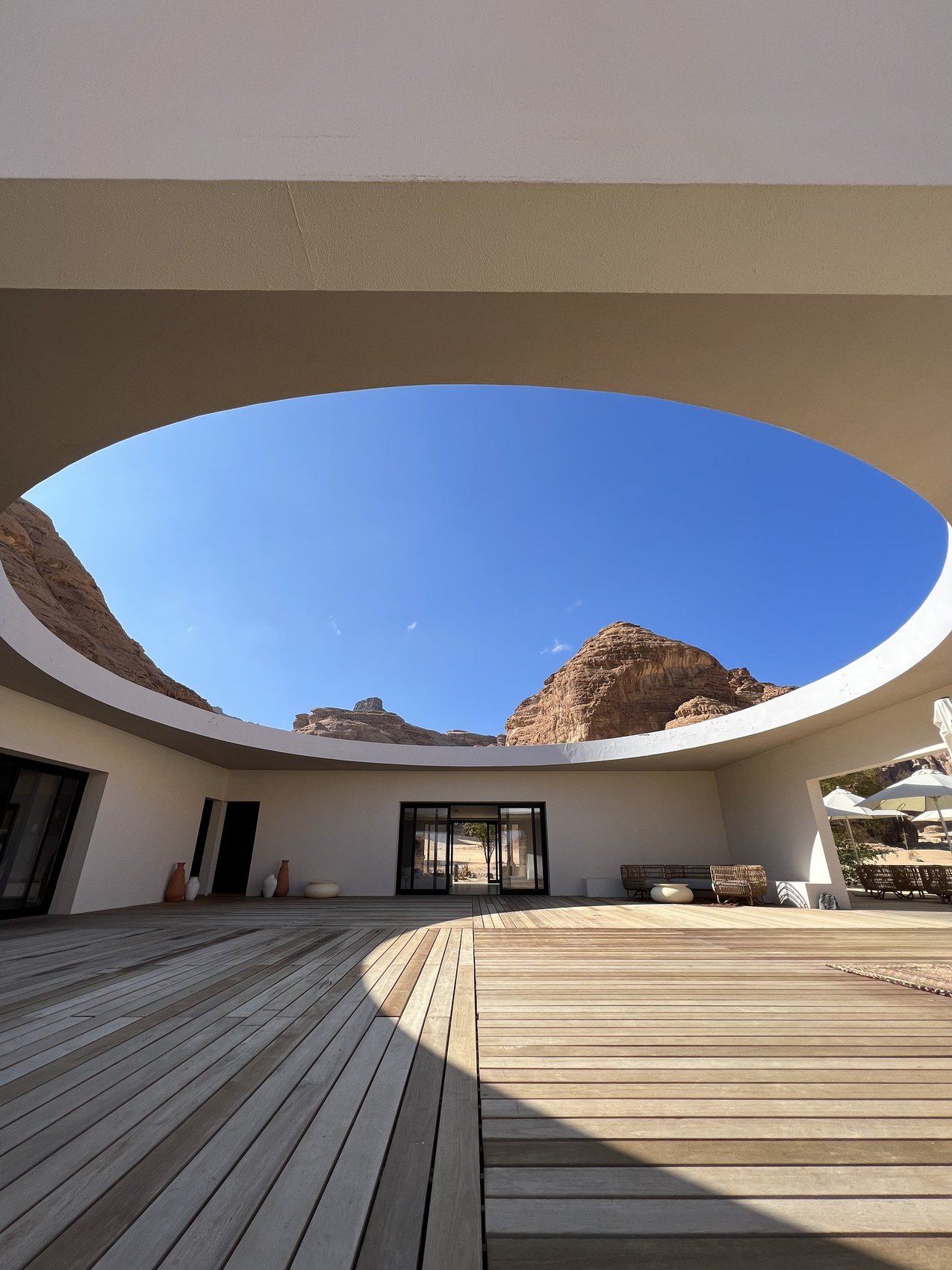 A round opening cut in a white roof at Habitas AlUla with the sandstone cliff visible through it above a timber deck