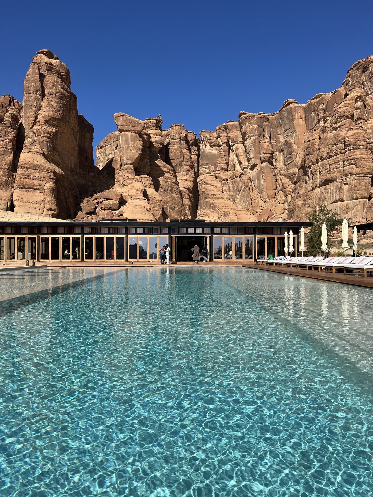 The long pool at Habitas AlUla in bright sun with the restaurant colonnade at the far end and sandstone cliffs behind