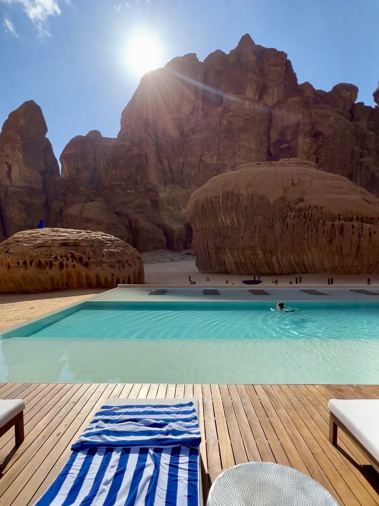 The pool at Habitas AlUla in full sun, a swimmer crossing it, with the sandstone formations of Ashar Valley rising straight out of the sand behind