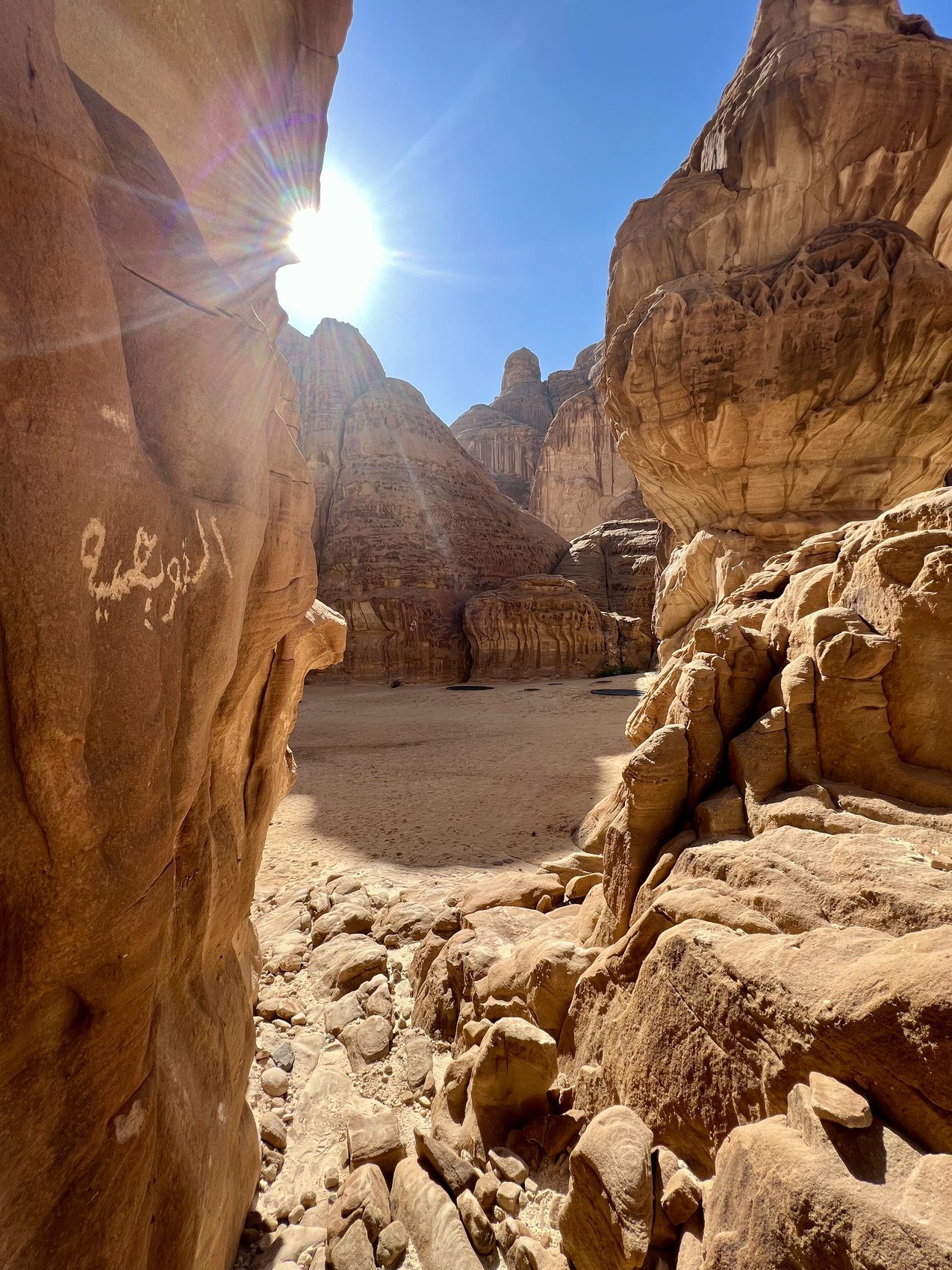 The sun flaring over the rim of a sandstone canyon at AlUla with Arabic script cut into the rock below