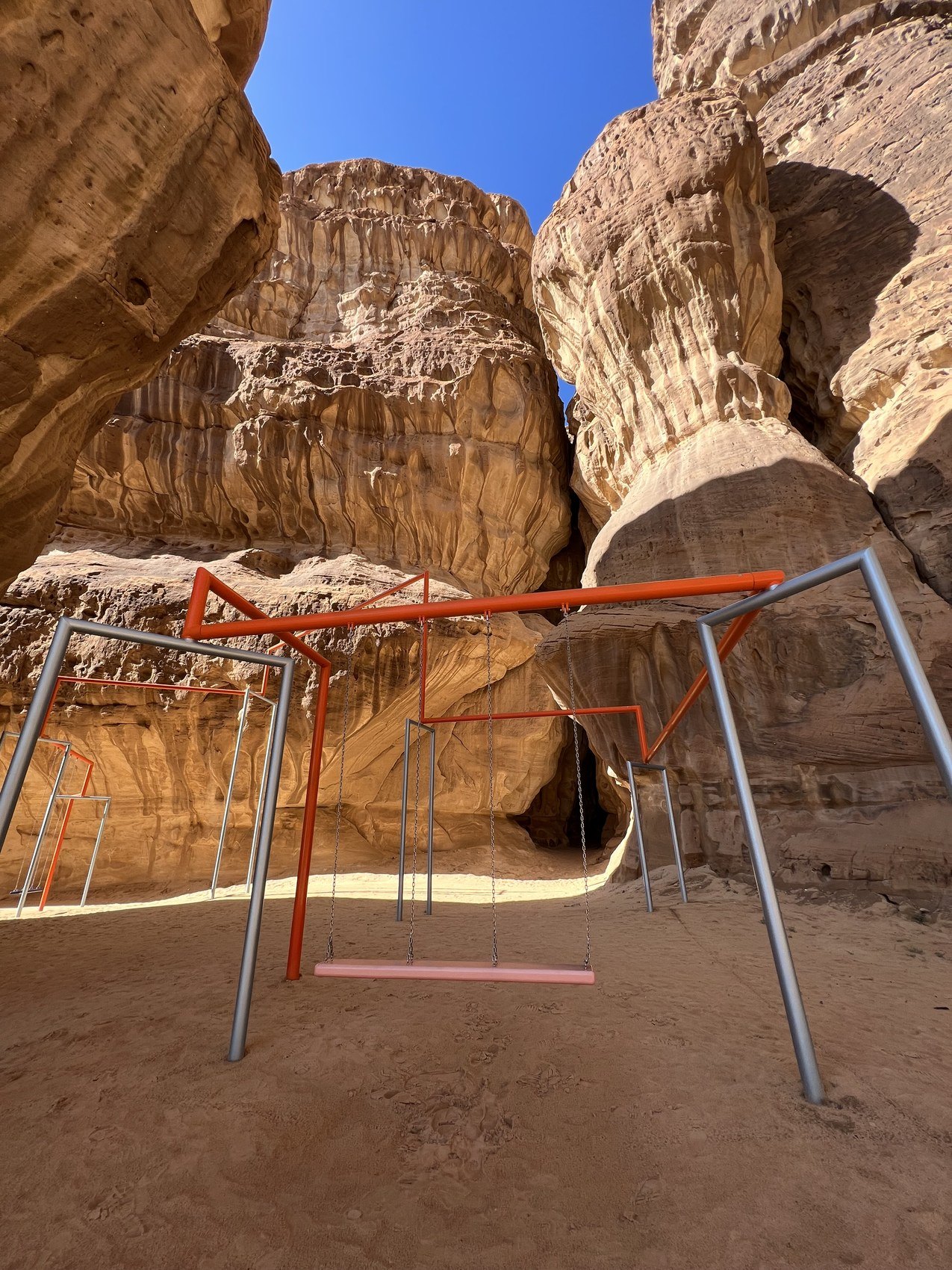 A red and grey metal swing frame standing on the sand between tall sandstone towers at AlUla