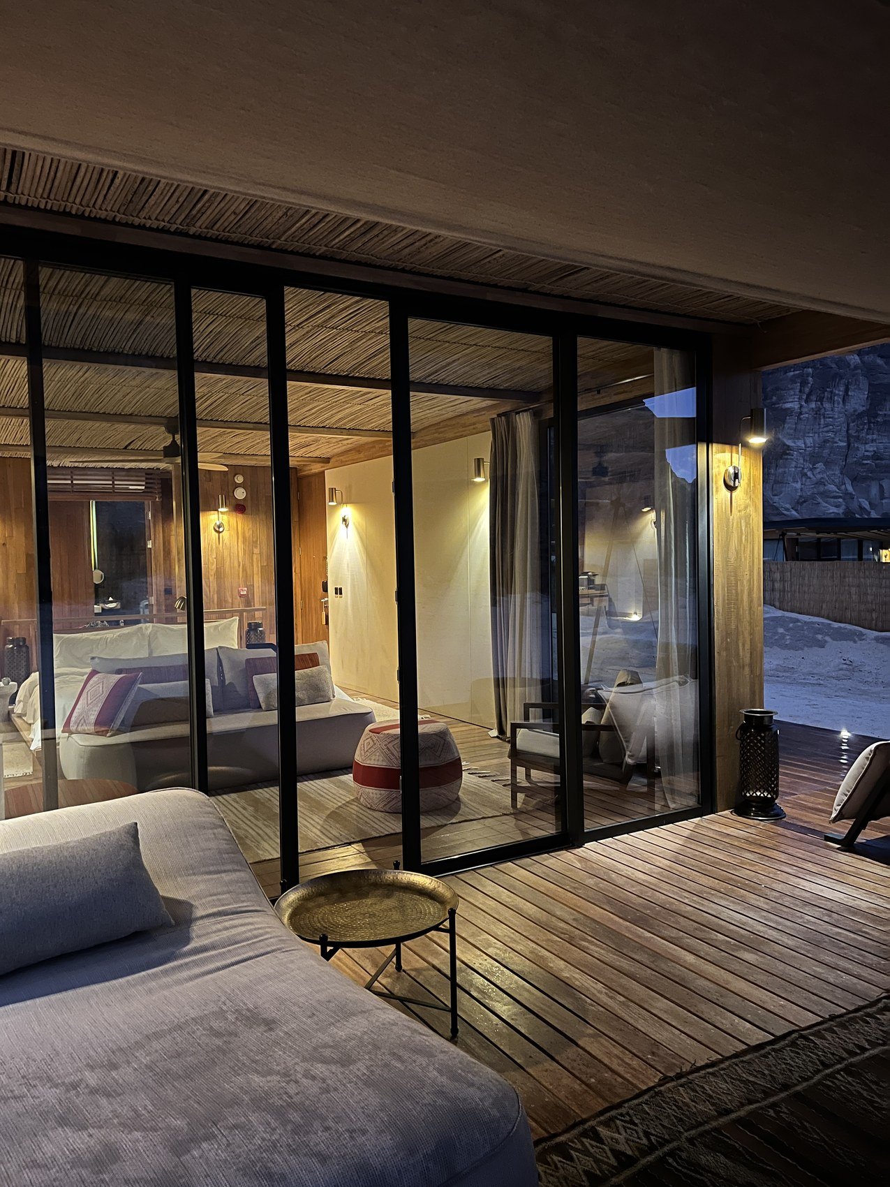 A tented villa at Habitas AlUla lit from within at dusk, its glass front open onto the terrace and the desert