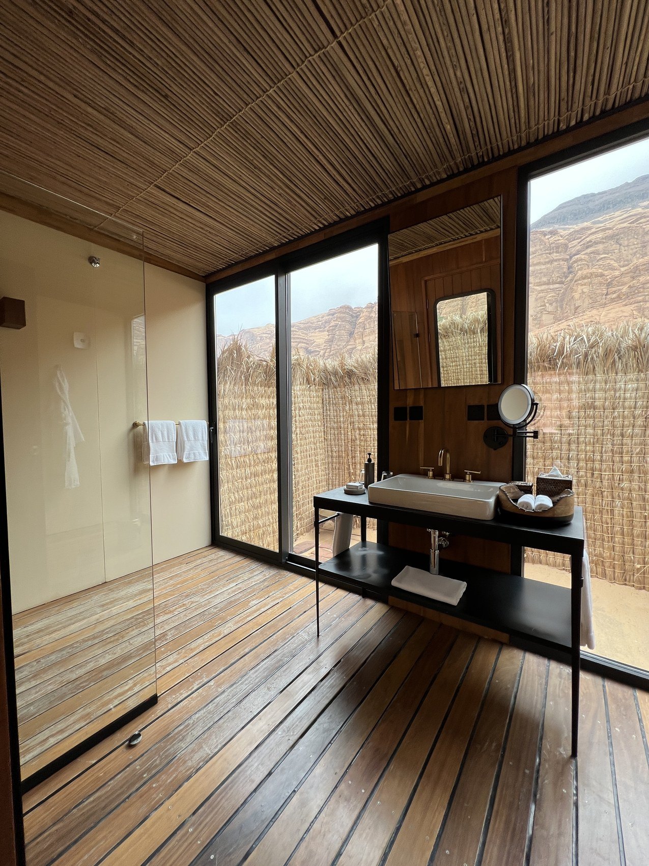 A bathroom at Habitas AlUla with a glass wall onto the canyon, a stone basin on a black frame and a reed ceiling