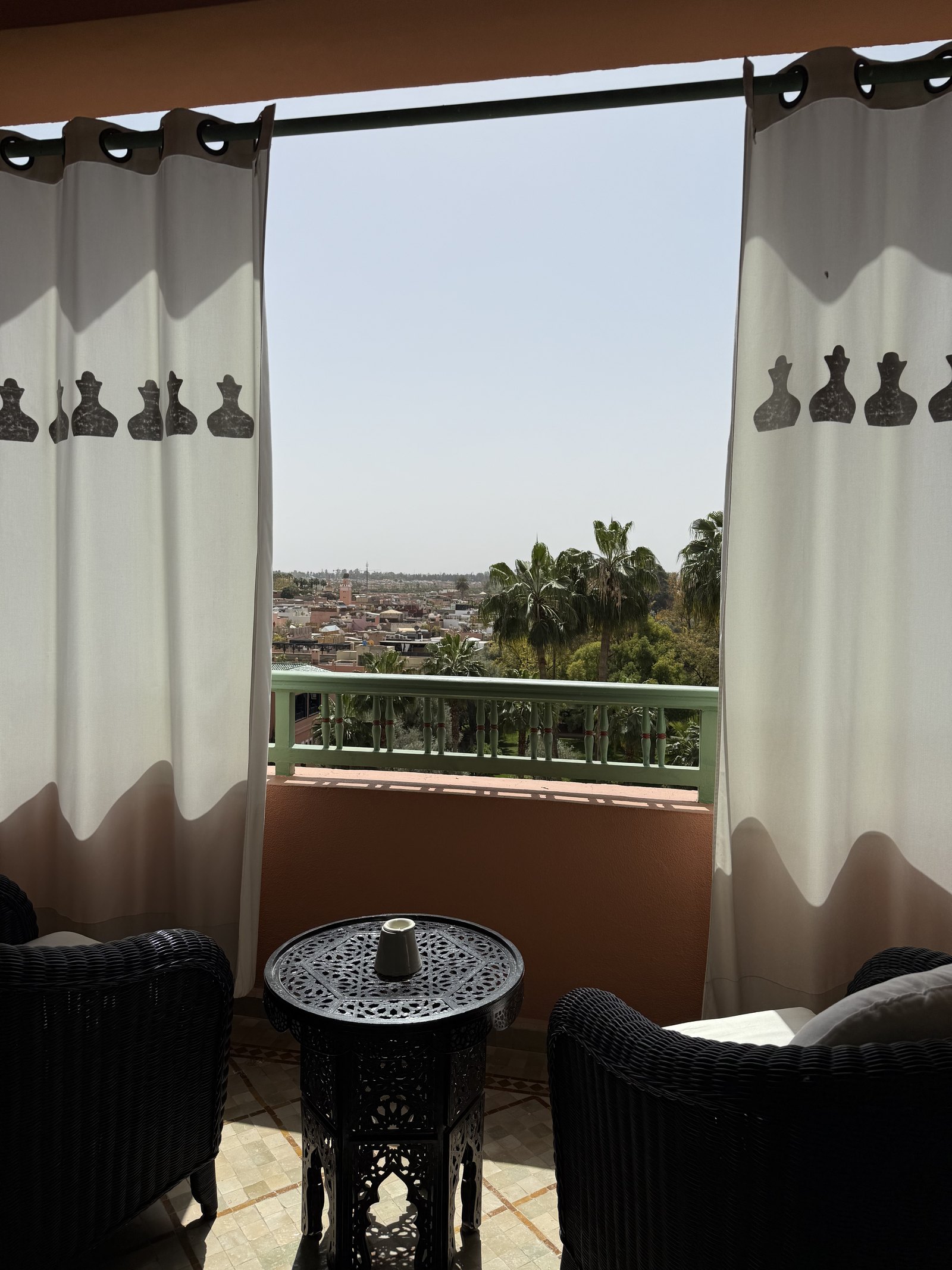 A balcony with white curtains opening onto the medina roofs and palms beyond