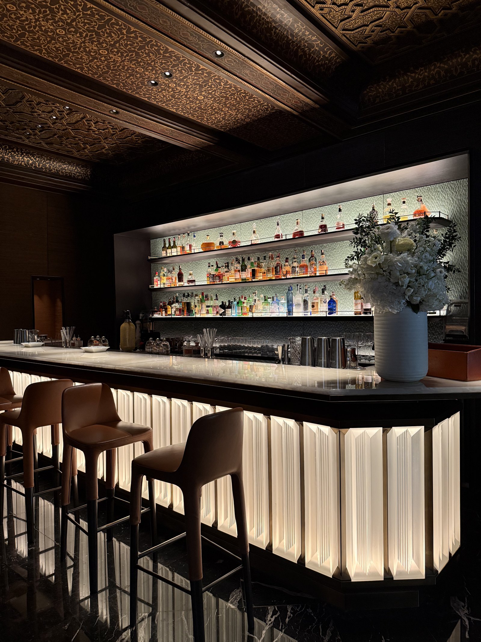 A bar with a lit back shelf of bottles, a white vase of flowers and tall leather stools