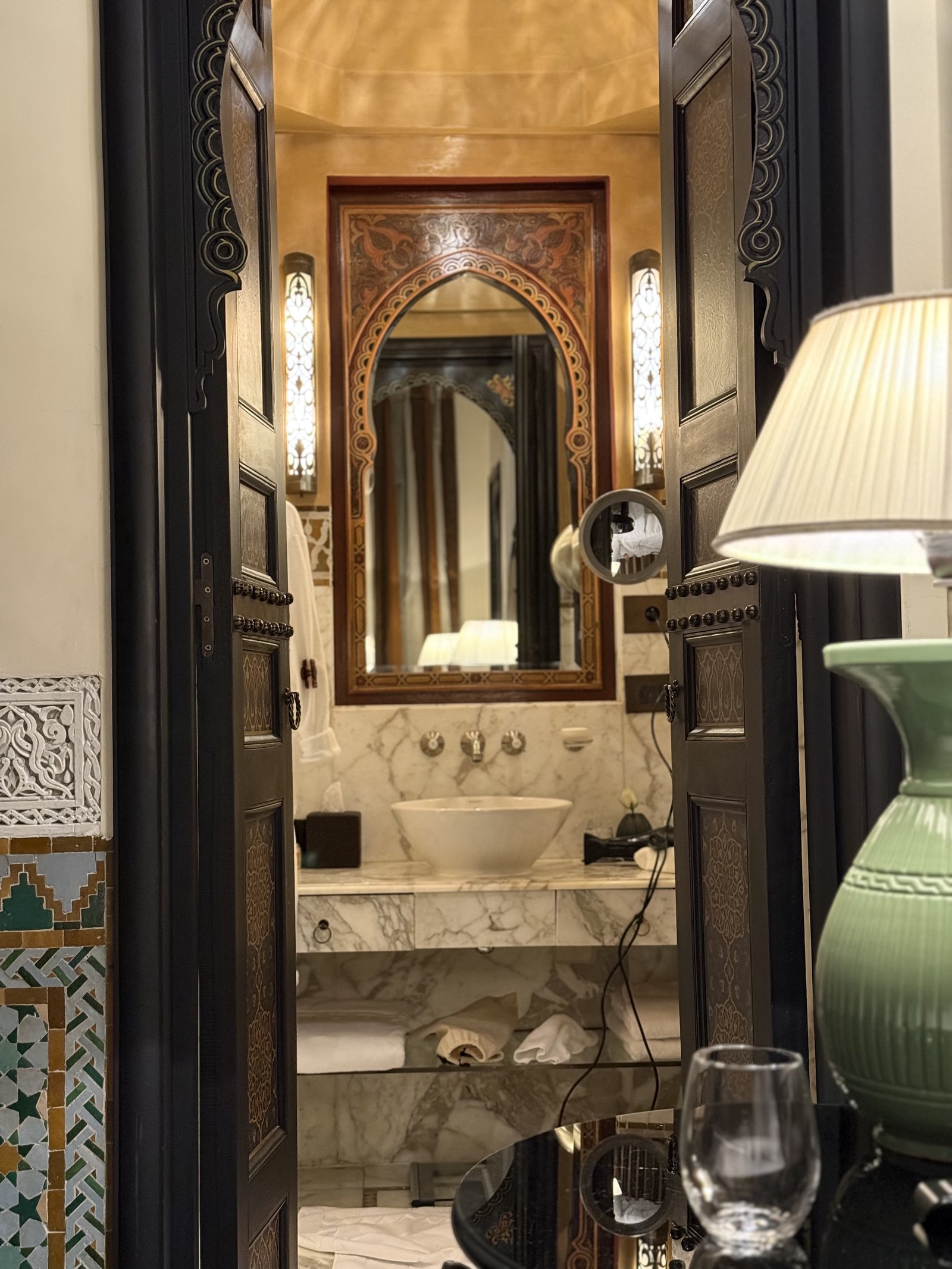 A carved wooden mirror above a marble basin, with a lamp and a glass on the shelf