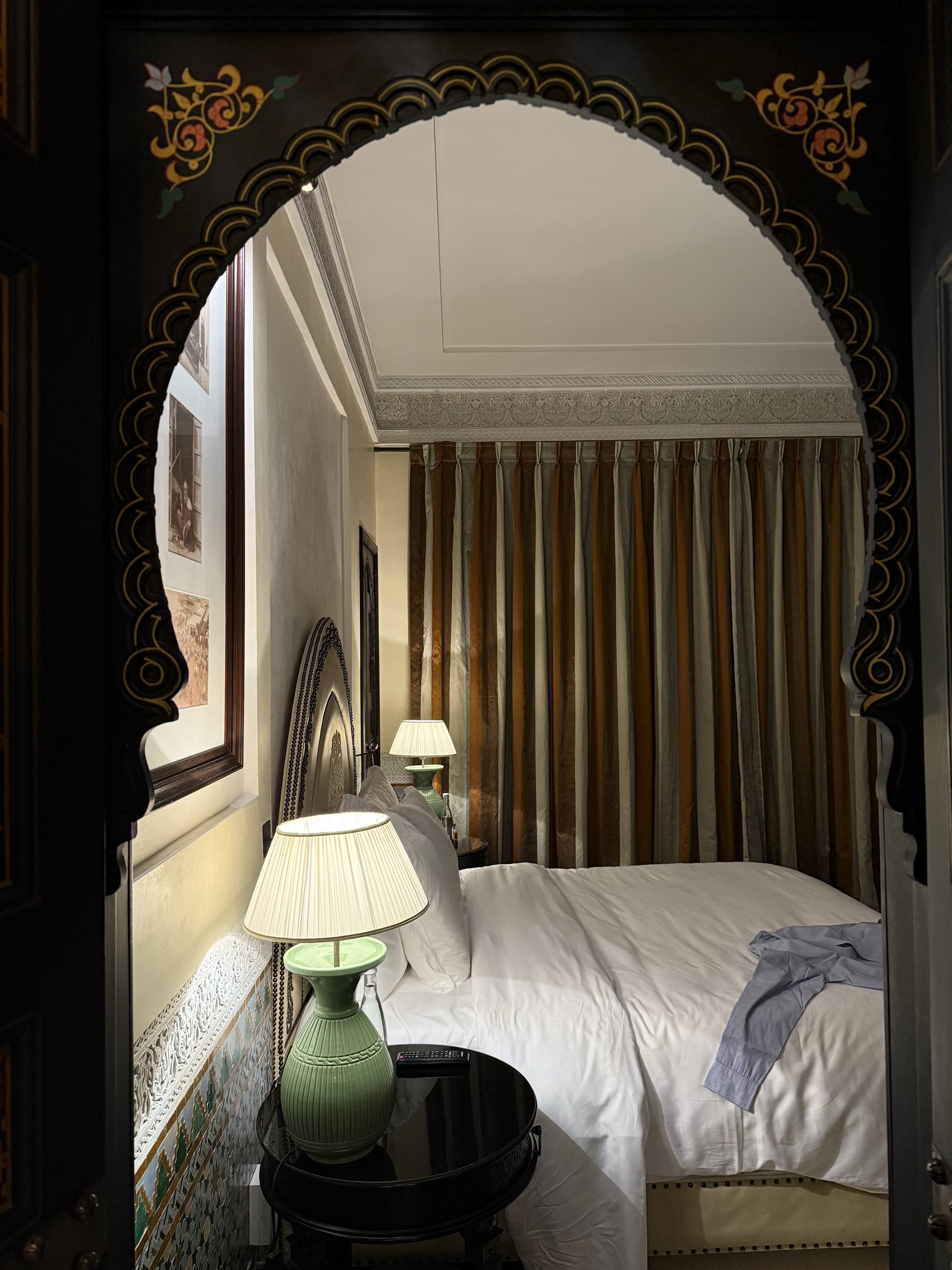 A bedroom seen through a carved wooden arch, with a bed, a green lamp and a striped curtain