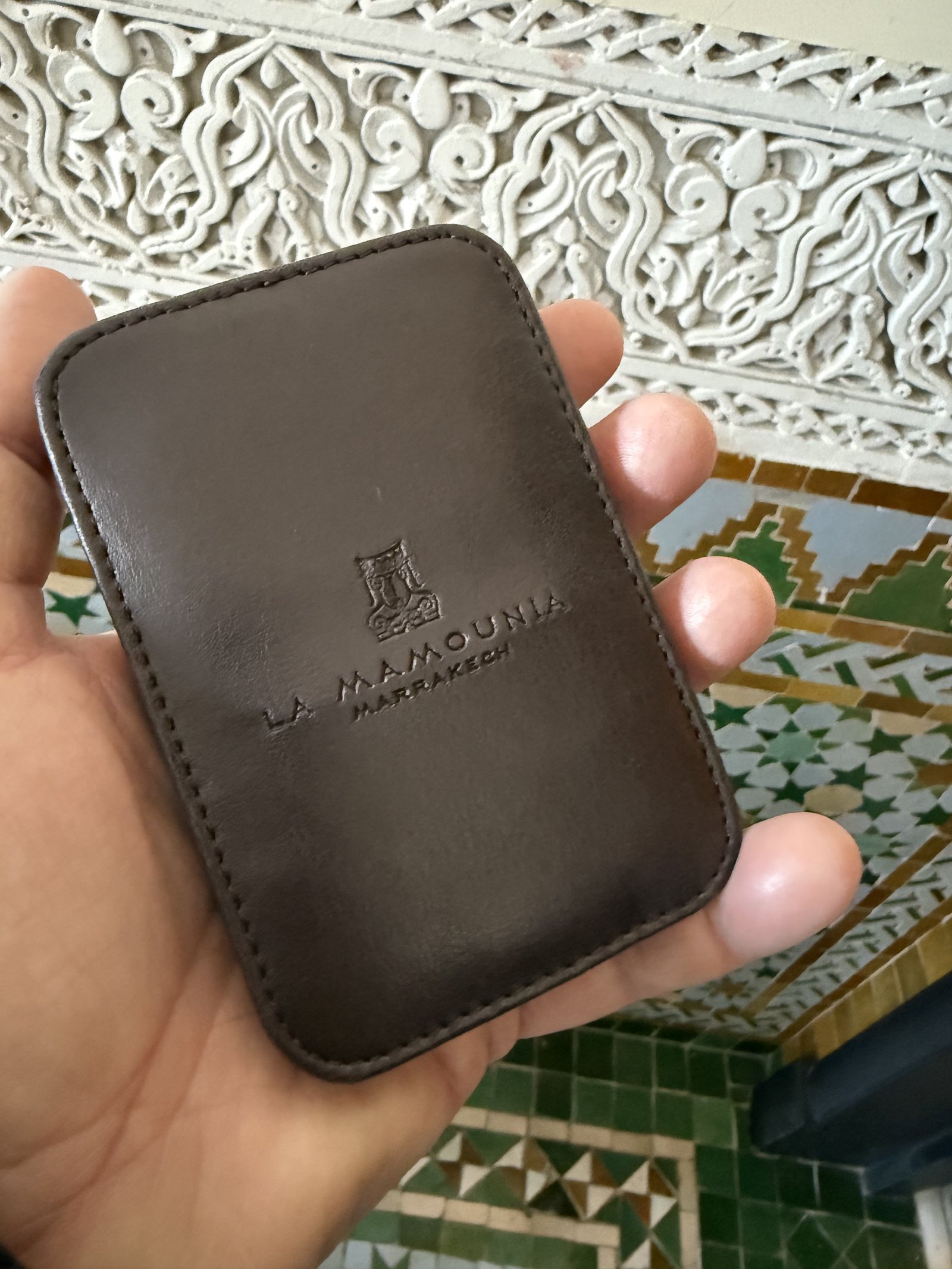 A dark brown leather card holder held in a hand, with the name La Mamounia Marrakech pressed into the cover above a carved plaster ceiling and zellige tiles