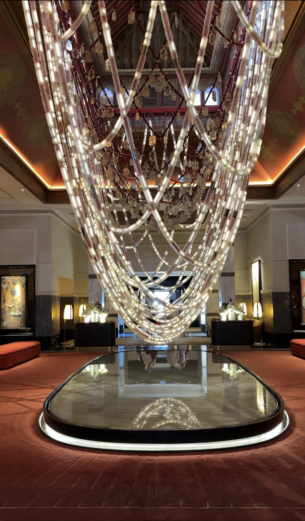 A chandelier of illuminated tubes strung in loops over a lounge, with a red painted ceiling above